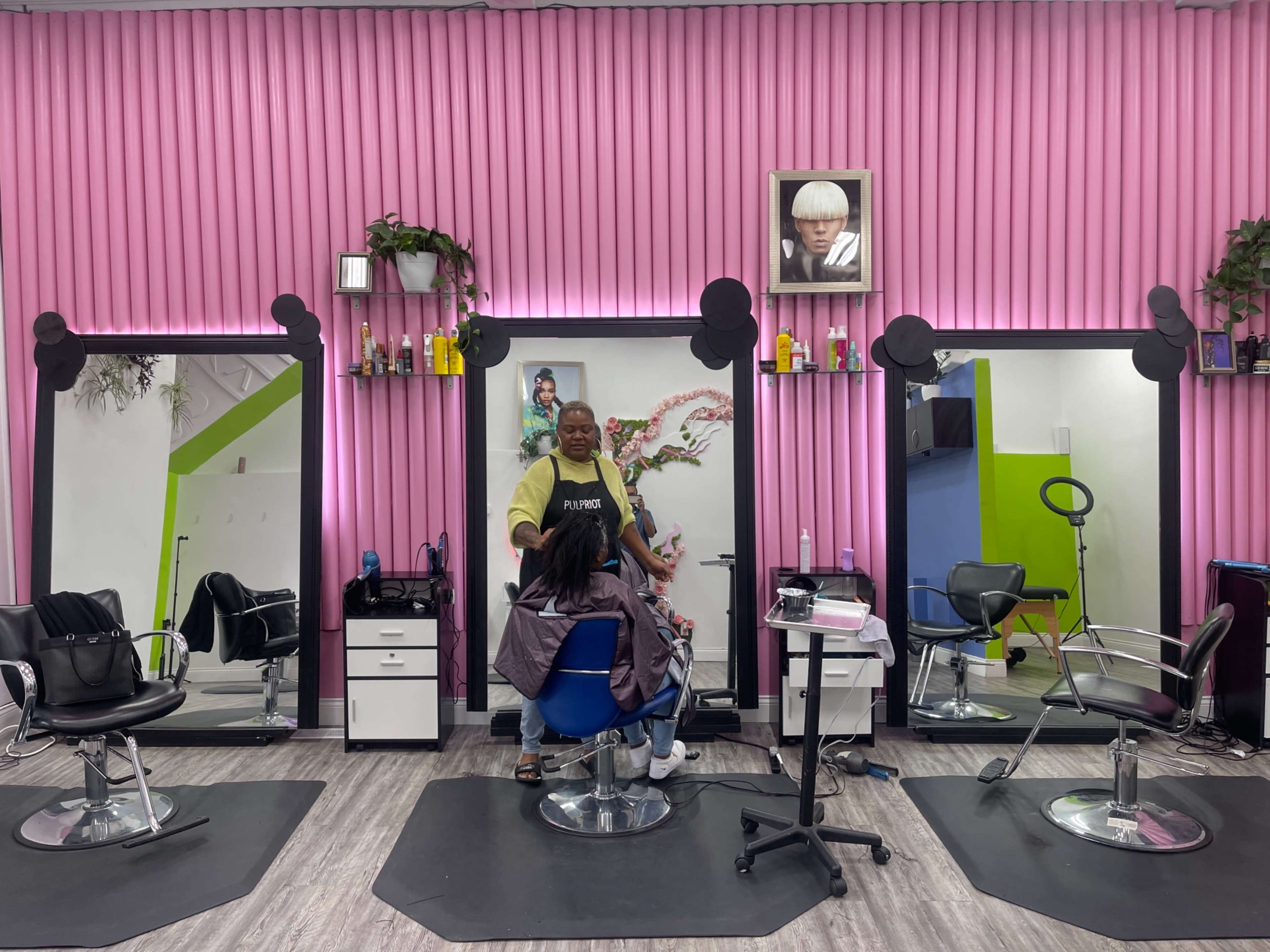 A hair salon features three styling stations with mirrors, pink walls, and a stylist working with a client in a chair.