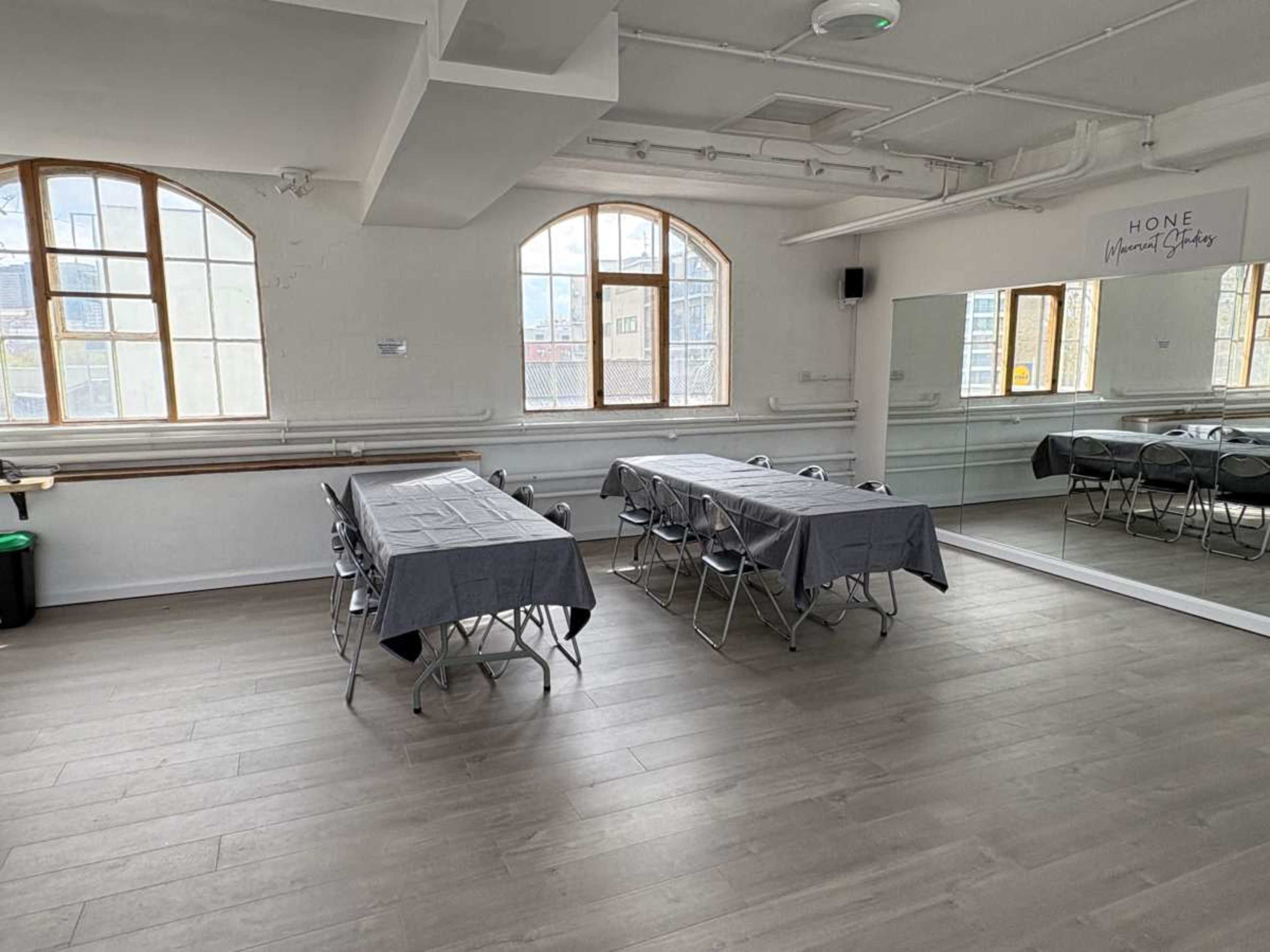 The image shows a spacious room with two long tables covered in black tablecloths and surrounded by folding chairs, with large windows and a mirror on one wall.