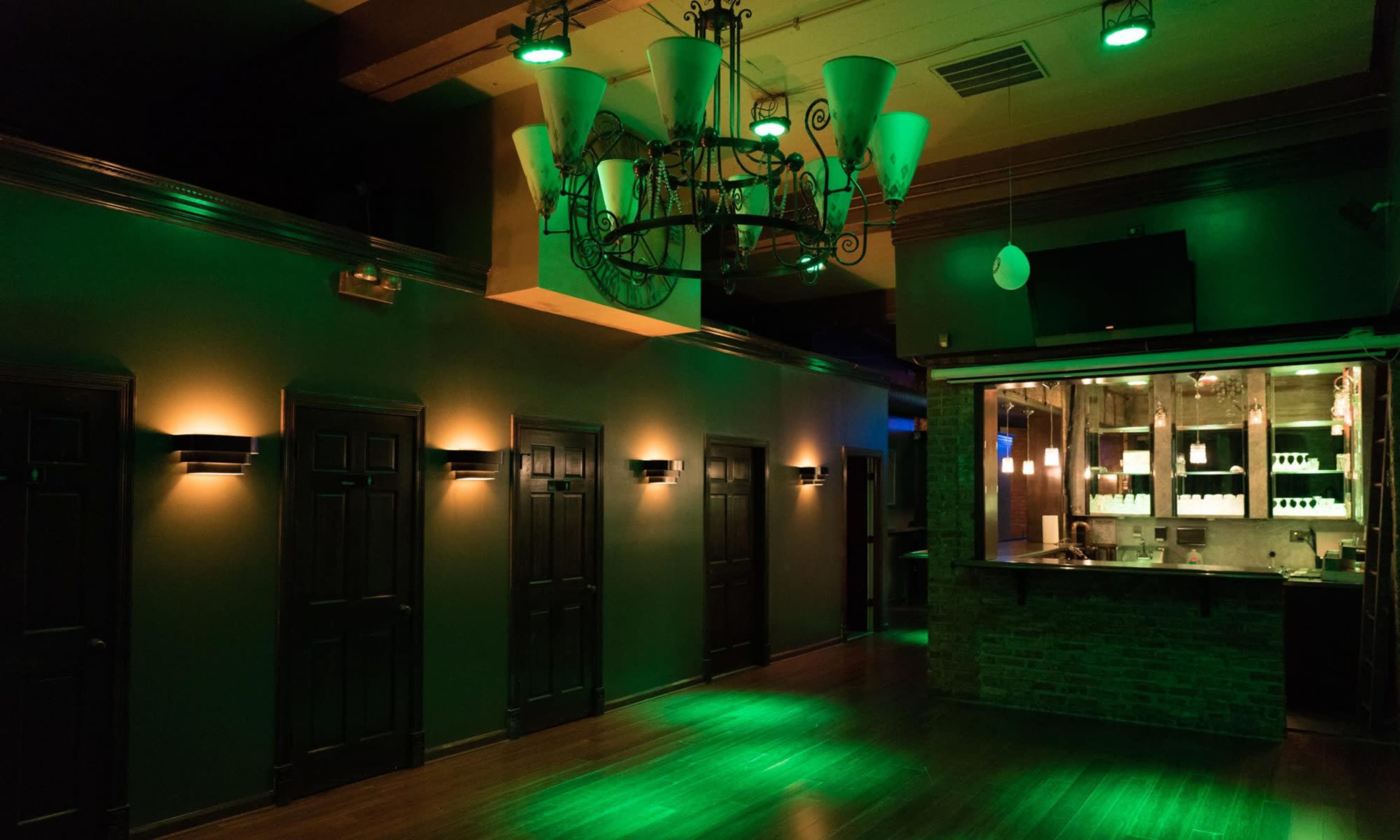 The image shows an empty dimly lit room with green lighting, featuring several dark wooden doors along the wall and a bar area in the background.