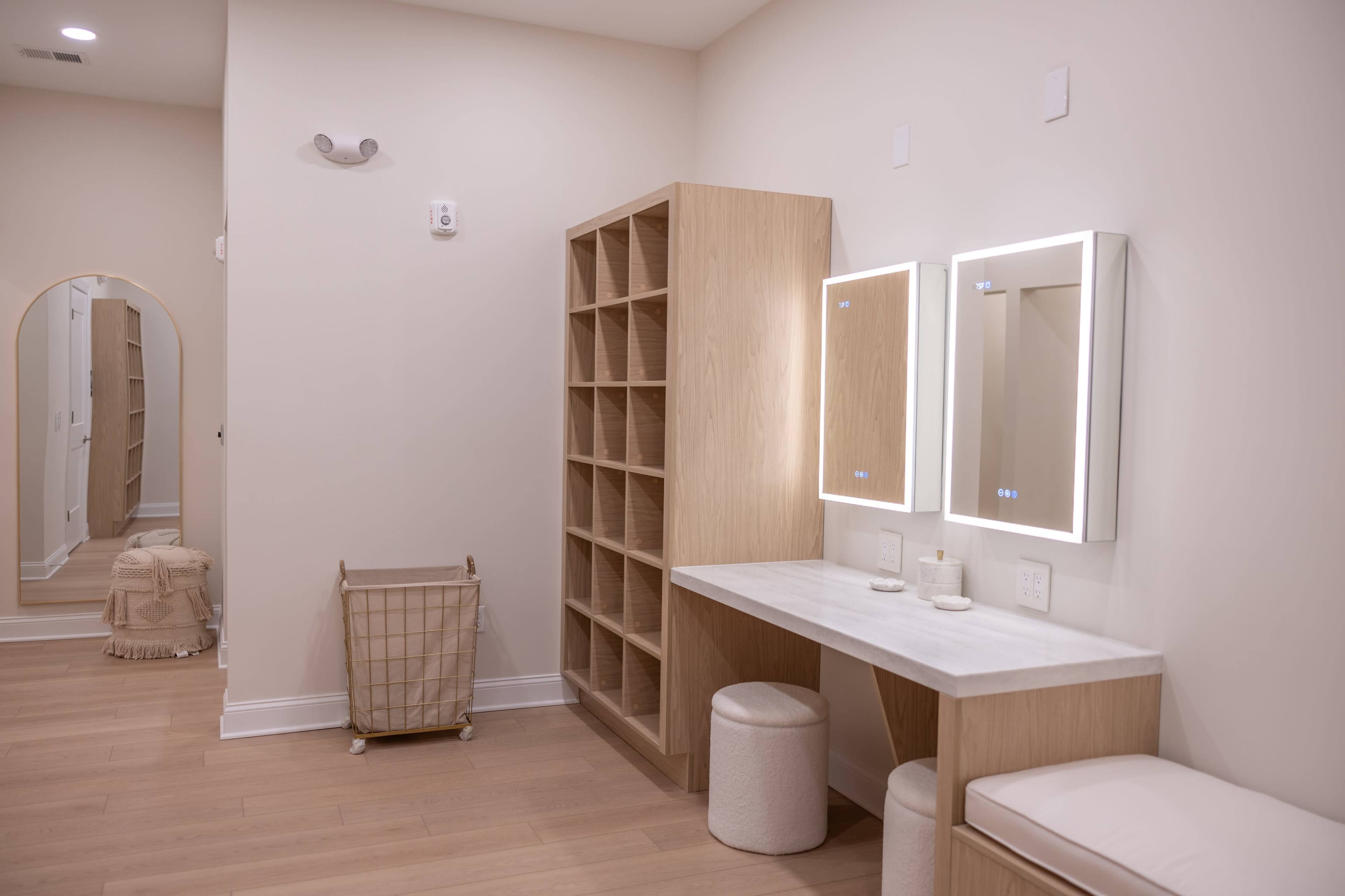 The image shows a minimalist room featuring a wooden shelf unit, a marble-topped vanity with illuminated mirrors, and a small storage cart in a neutral-colored environment.