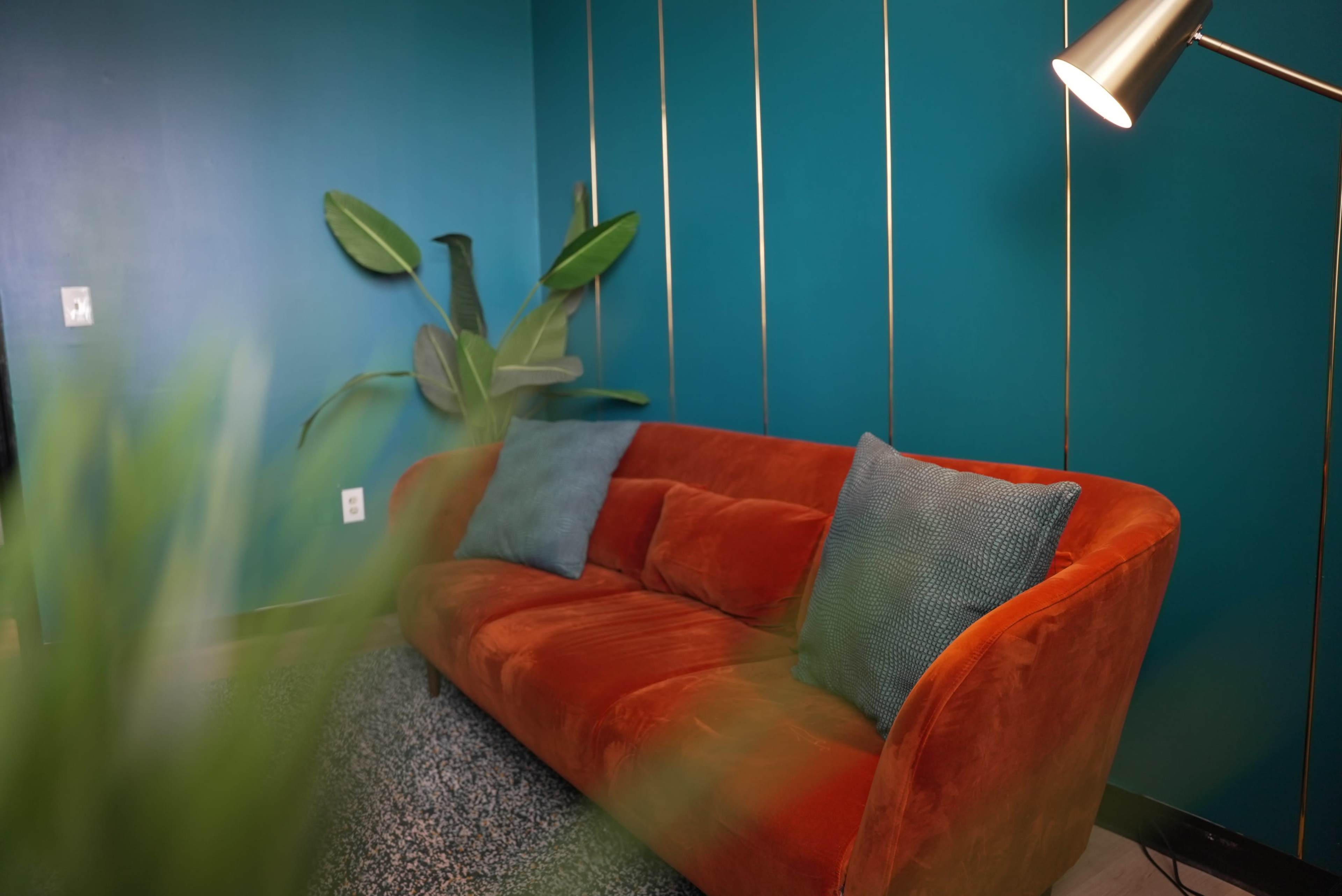 The image shows a vibrant teal wall with a sleek orange sofa adorned with two blue pillows, complemented by a green plant and a standing lamp.