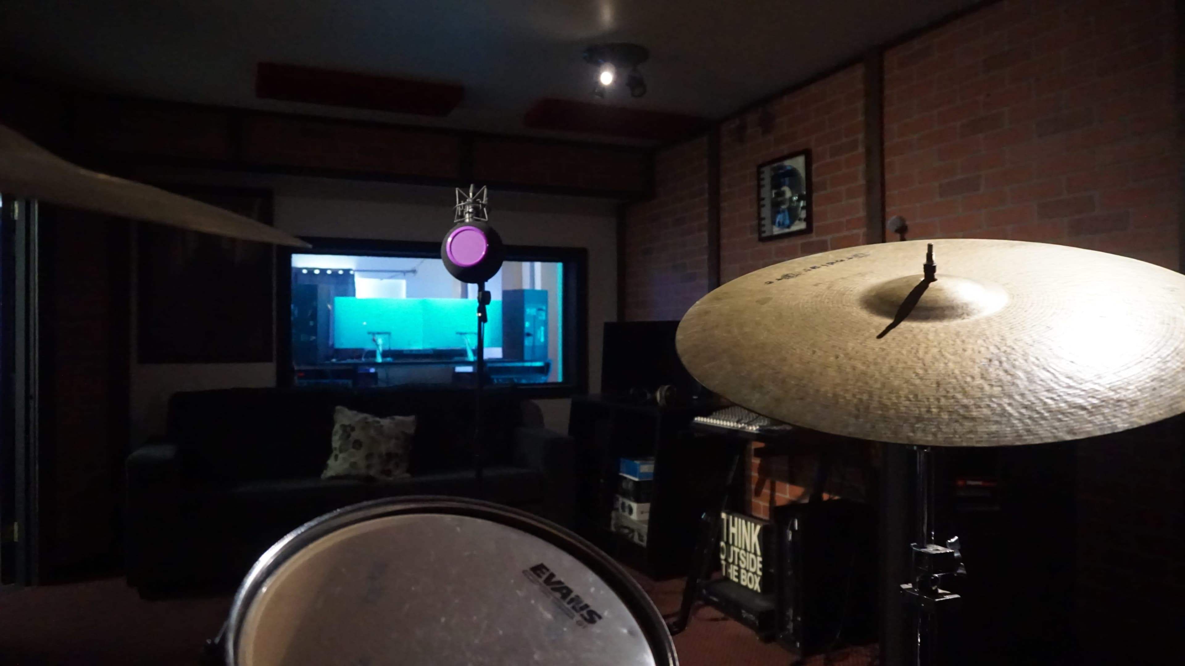 The image shows a recording studio with a drum set in the foreground, facing a soundproof booth that contains audio equipment, visible through a glass window.