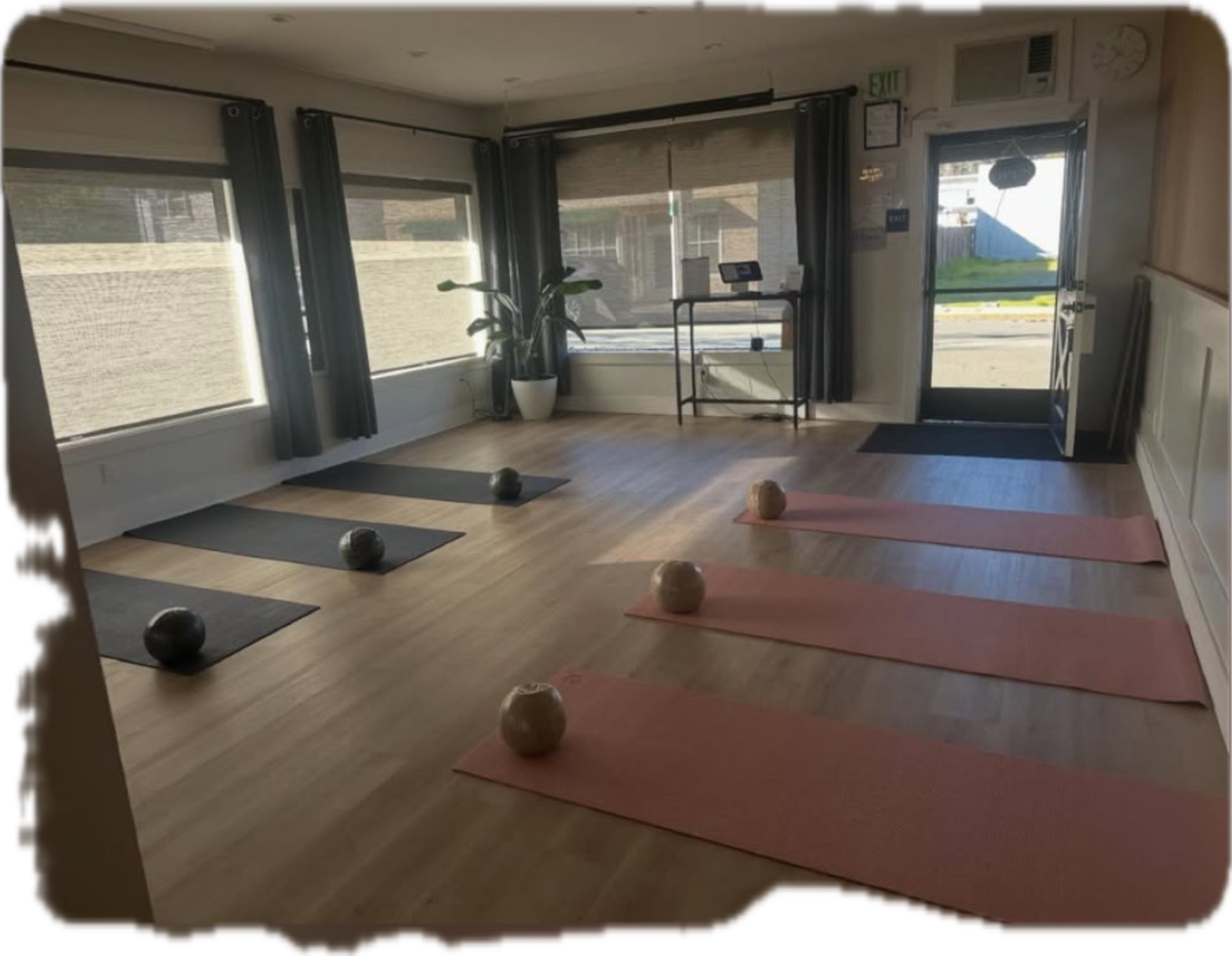 The image shows a fitness studio with six exercise mats arranged on the floor, a plant in the corner, and large windows allowing natural light to enter.