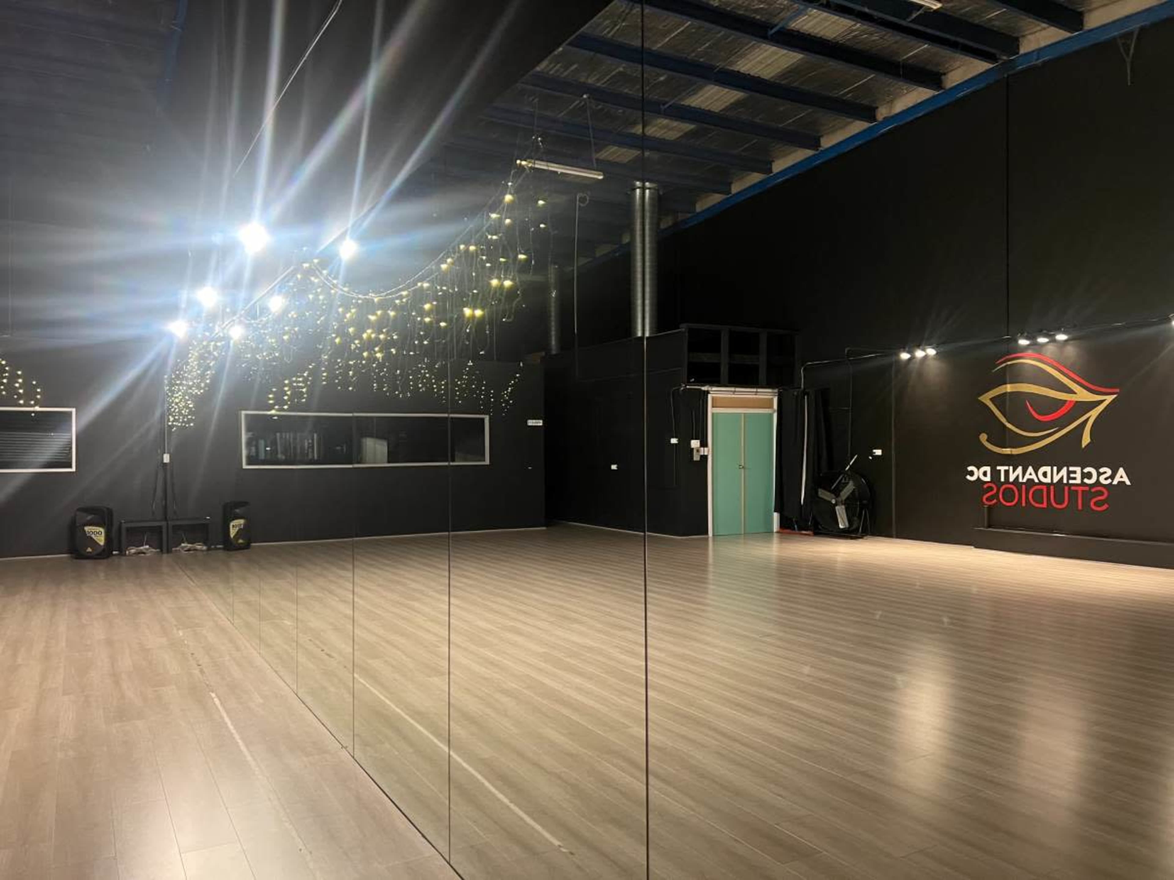 The image shows a spacious dance studio with wooden flooring, mirrored walls, and soft lighting, featuring the name "ASSOCIATED GC STUDIOS" displayed on the wall.