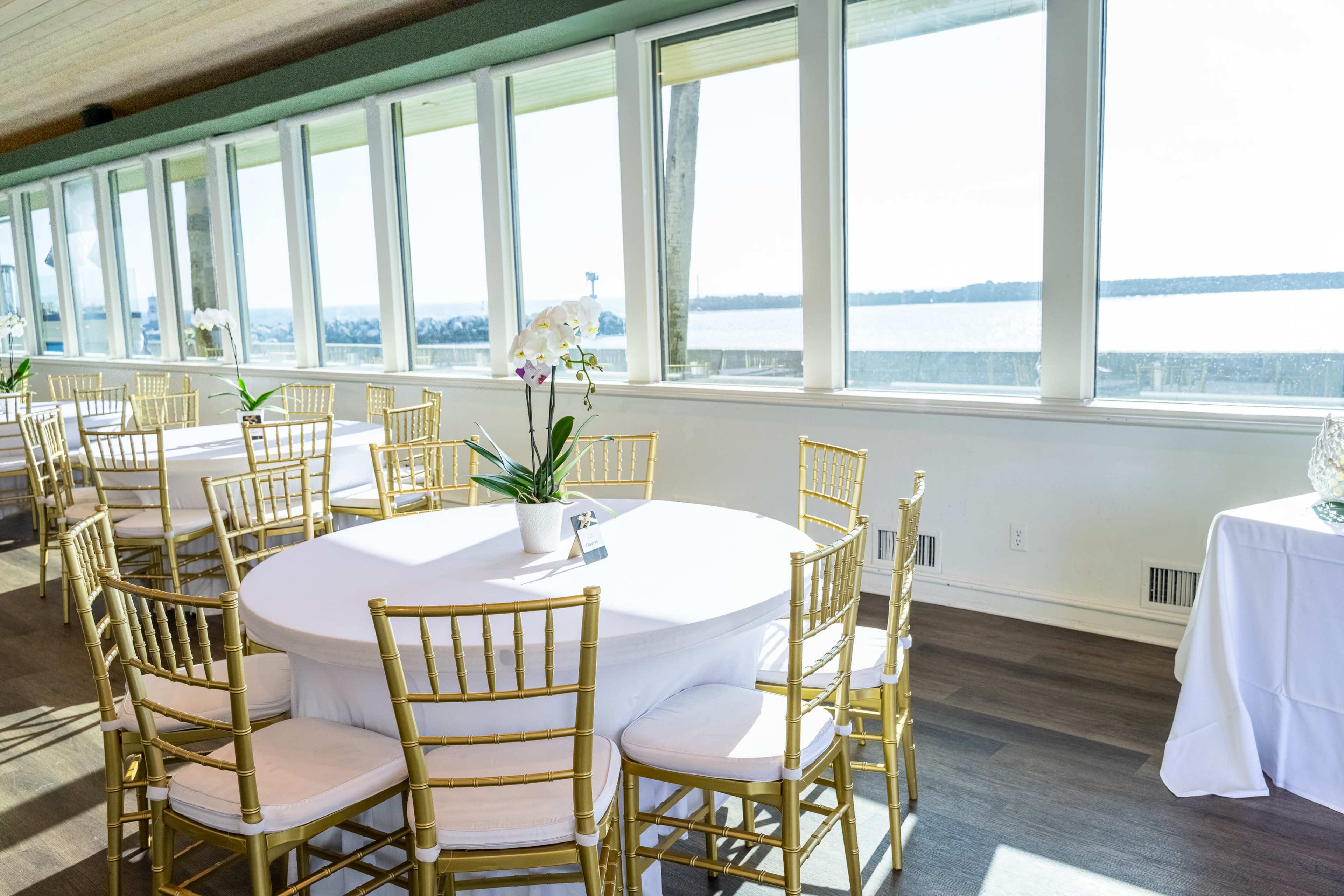 The scene shows a bright, spacious indoor venue with round tables set with white tablecloths and gold chairs, overlooking a waterfront through large windows.