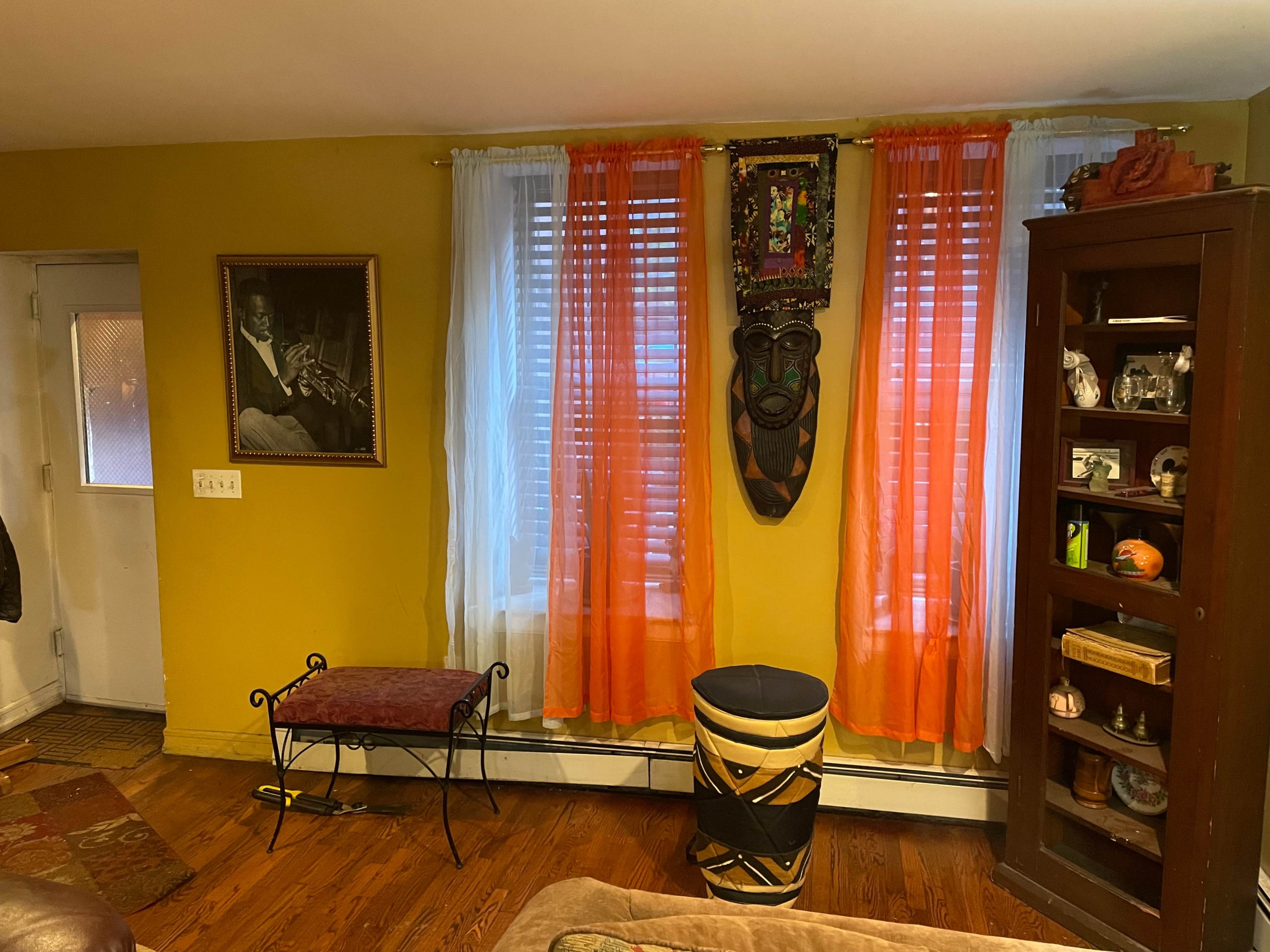 The image shows a living room with yellow walls, orange and white curtains, a wooden shelf displaying decorative items, and a framed picture on the wall.