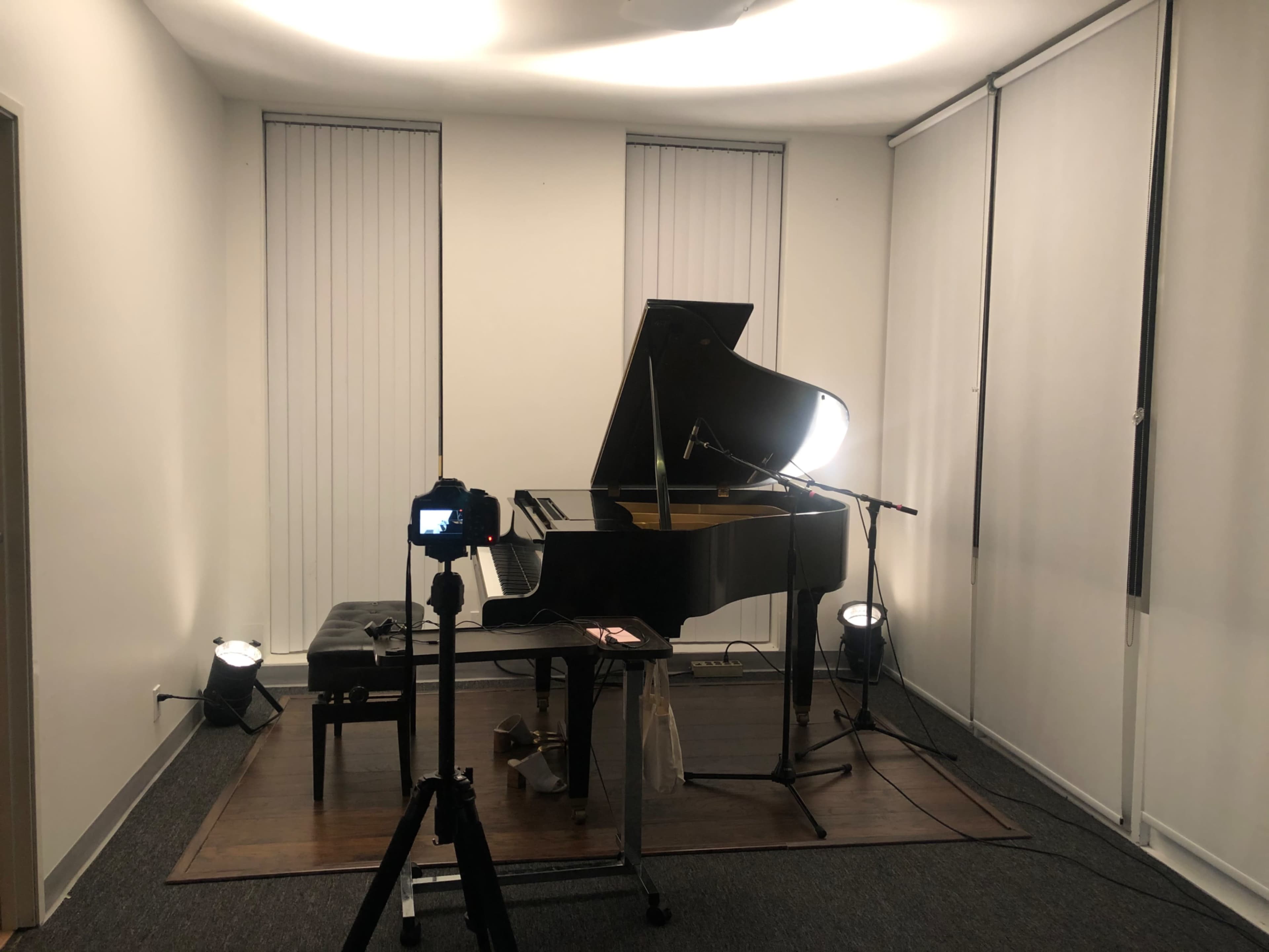 A grand piano set up in a minimalist room with soft lighting, a camera on a tripod positioned in front of it, and a microphone nearby.