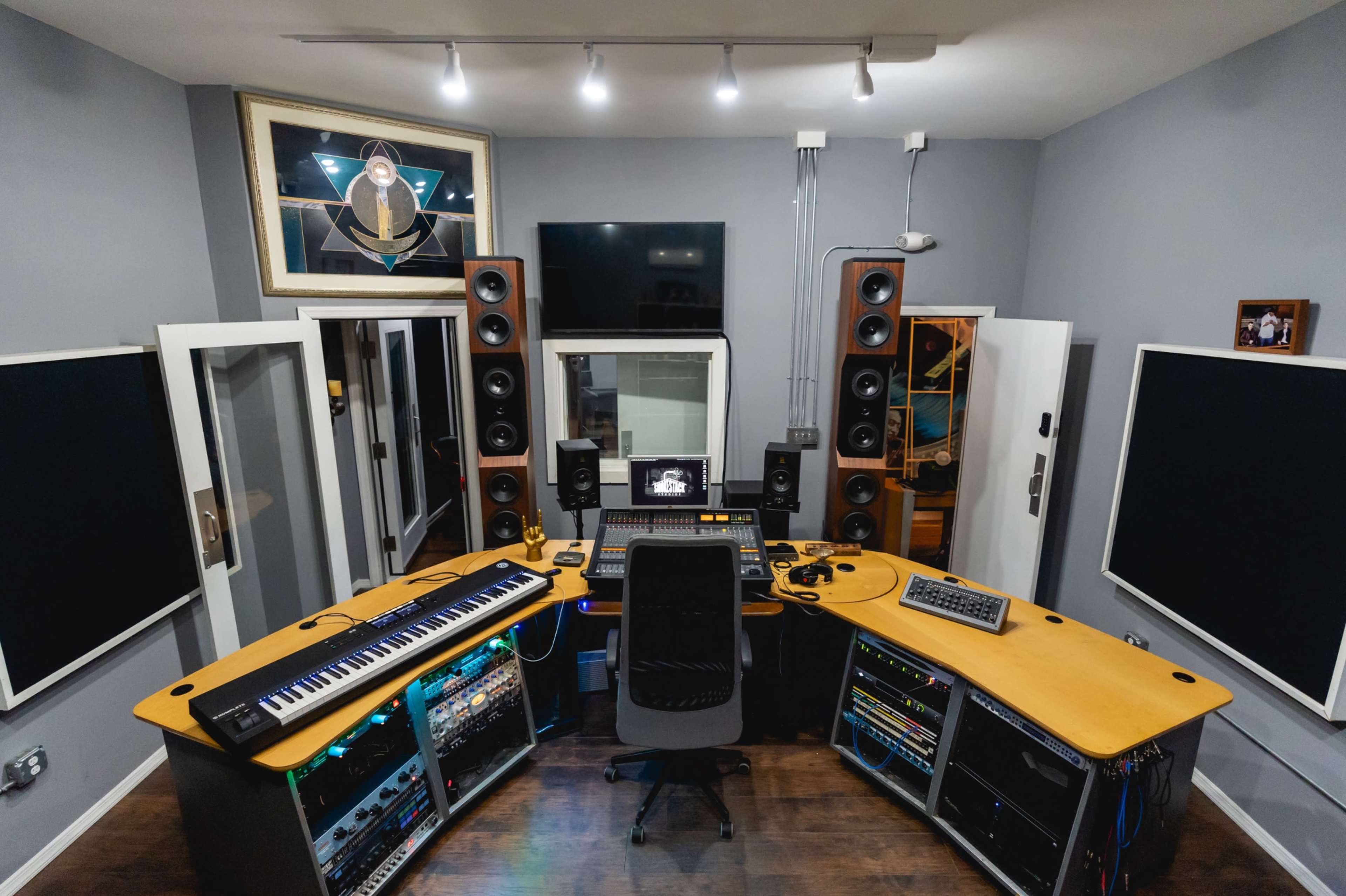 The image shows a modern recording studio with a curved desk containing audio equipment, speakers, and a keyboard, surrounded by soundproof panels and a large monitor.