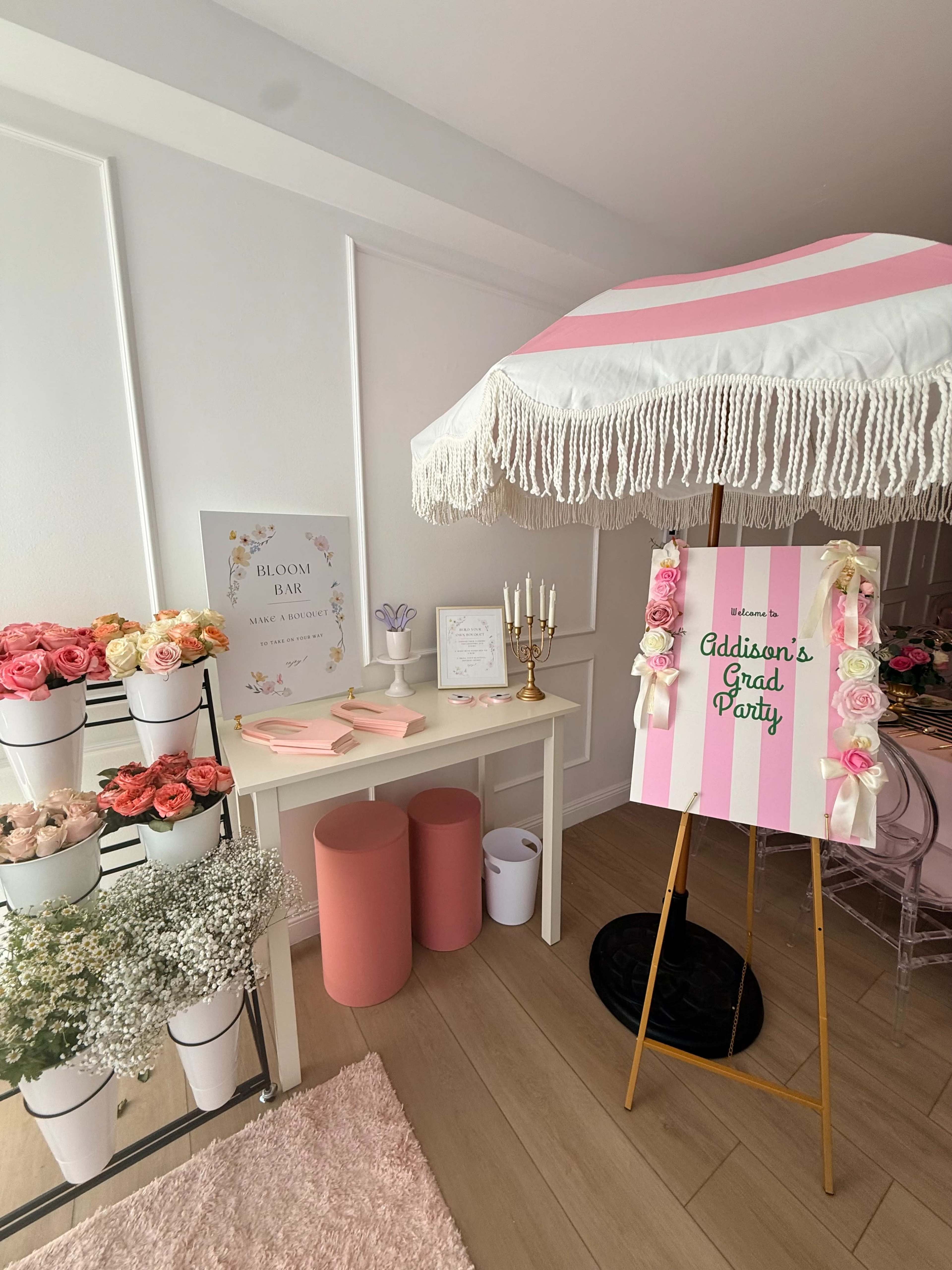 The image shows a decorated room featuring a floral display with pink and white roses, a sign for a graduation party, and an umbrella over a table with party supplies.