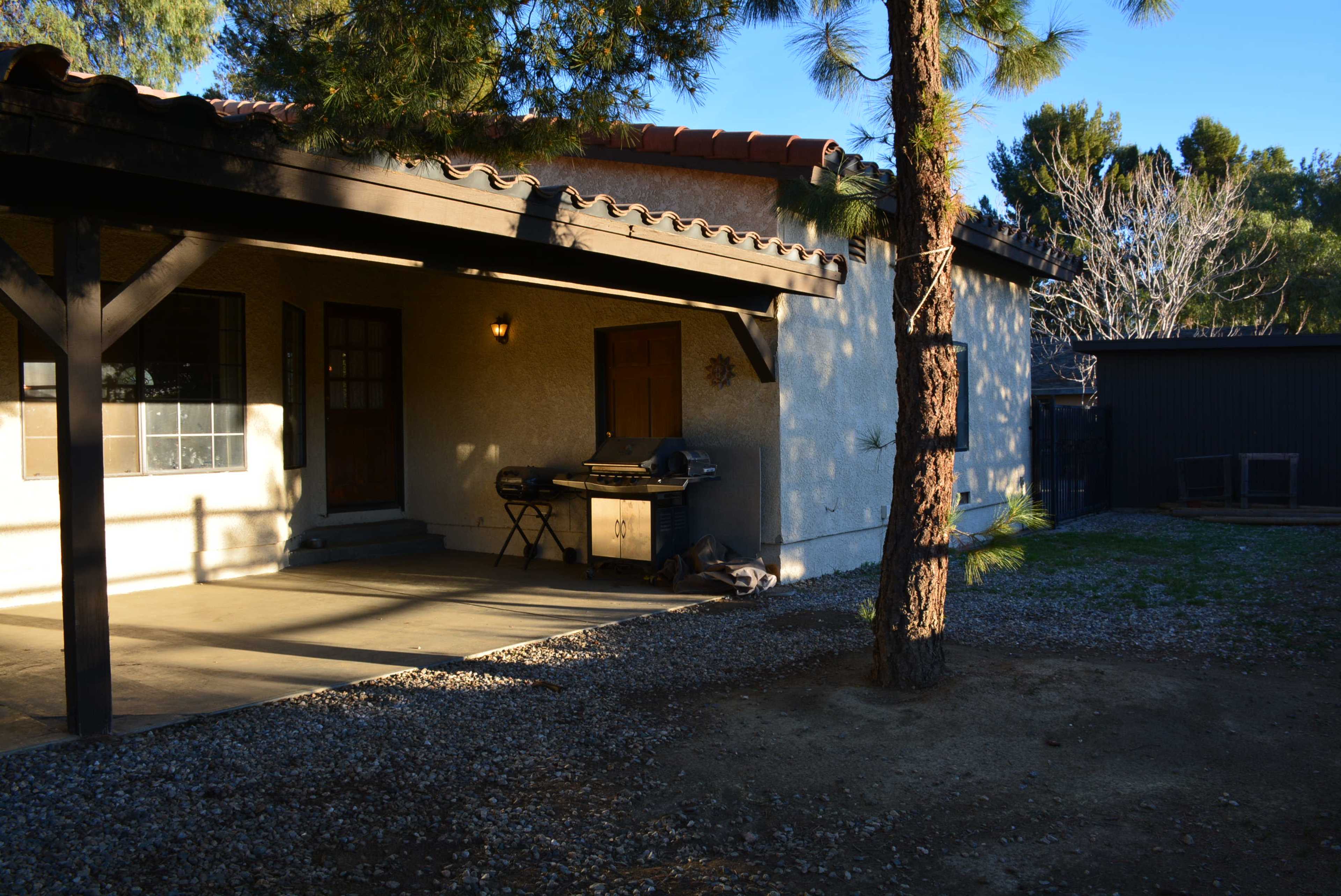 The image shows a patio area of a house featuring a grill and chair beside a tree, with gravel ground and a view of a fenced yard.