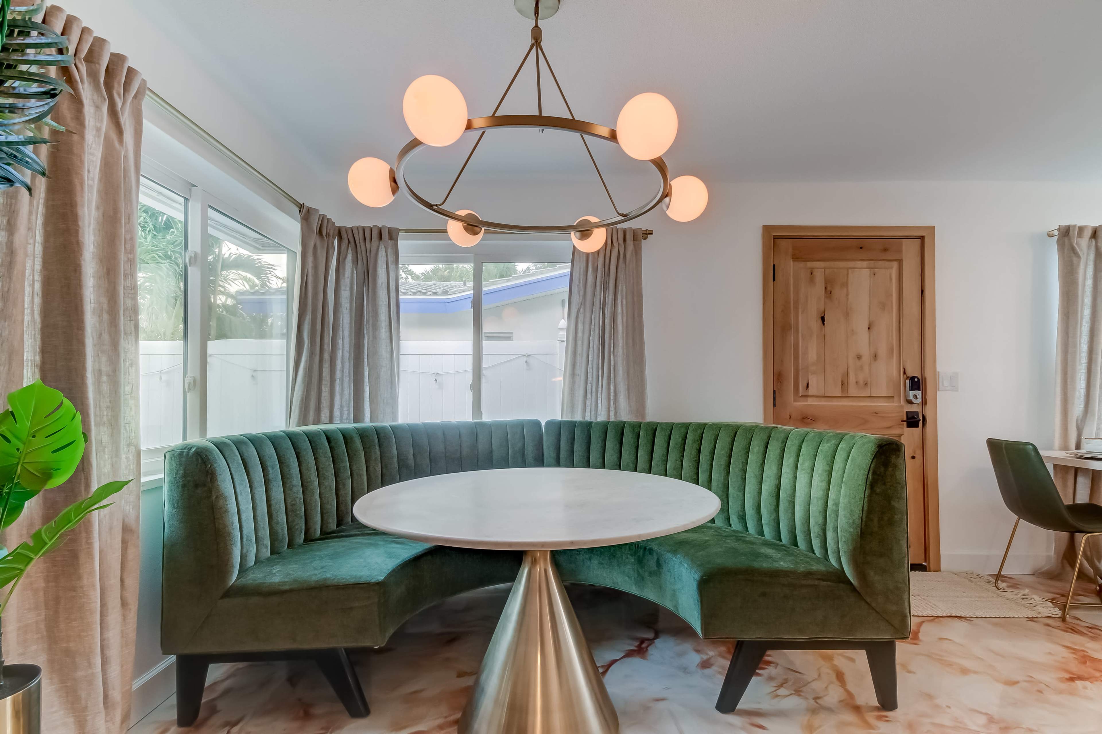 A modern dining area with a round table and a green upholstered semicircle bench, surrounded by windows and featuring a decorative light fixture.