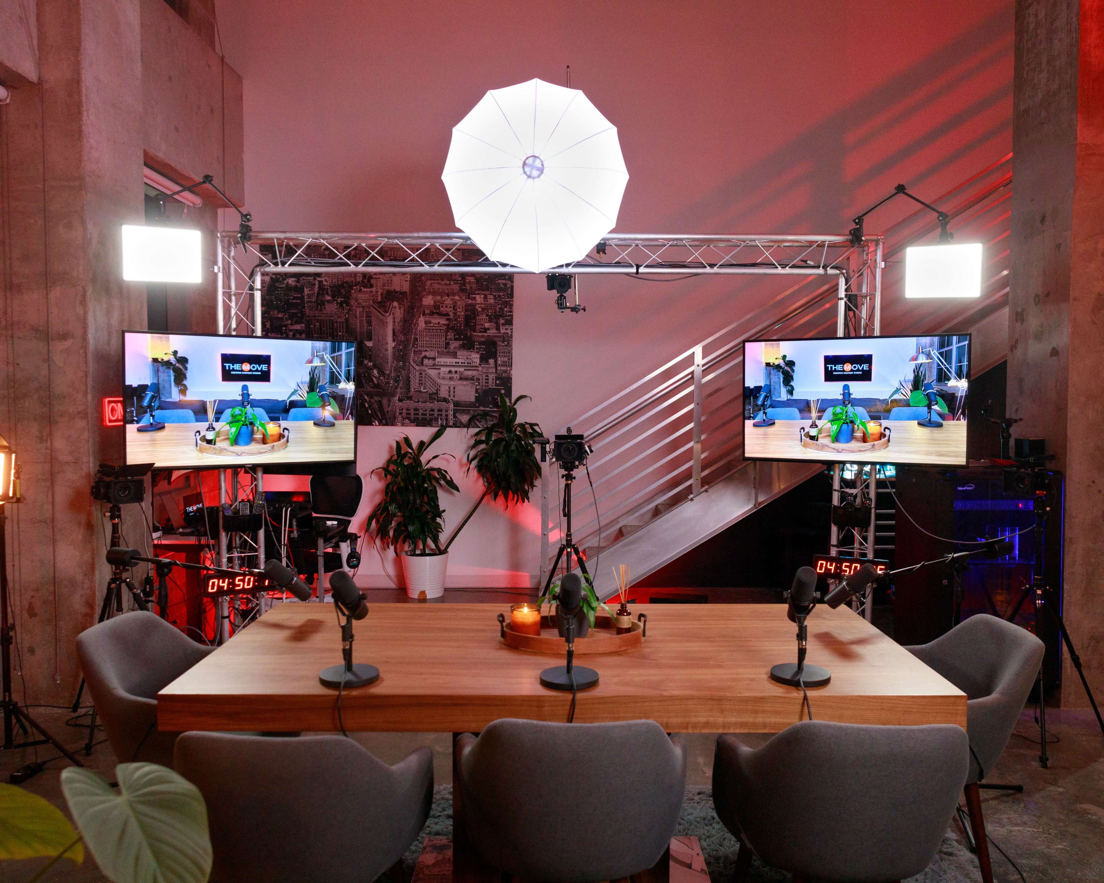 A modern studio setup featuring a large wooden table surrounded by chairs, two monitors displaying a presentation, and professional lighting equipment.