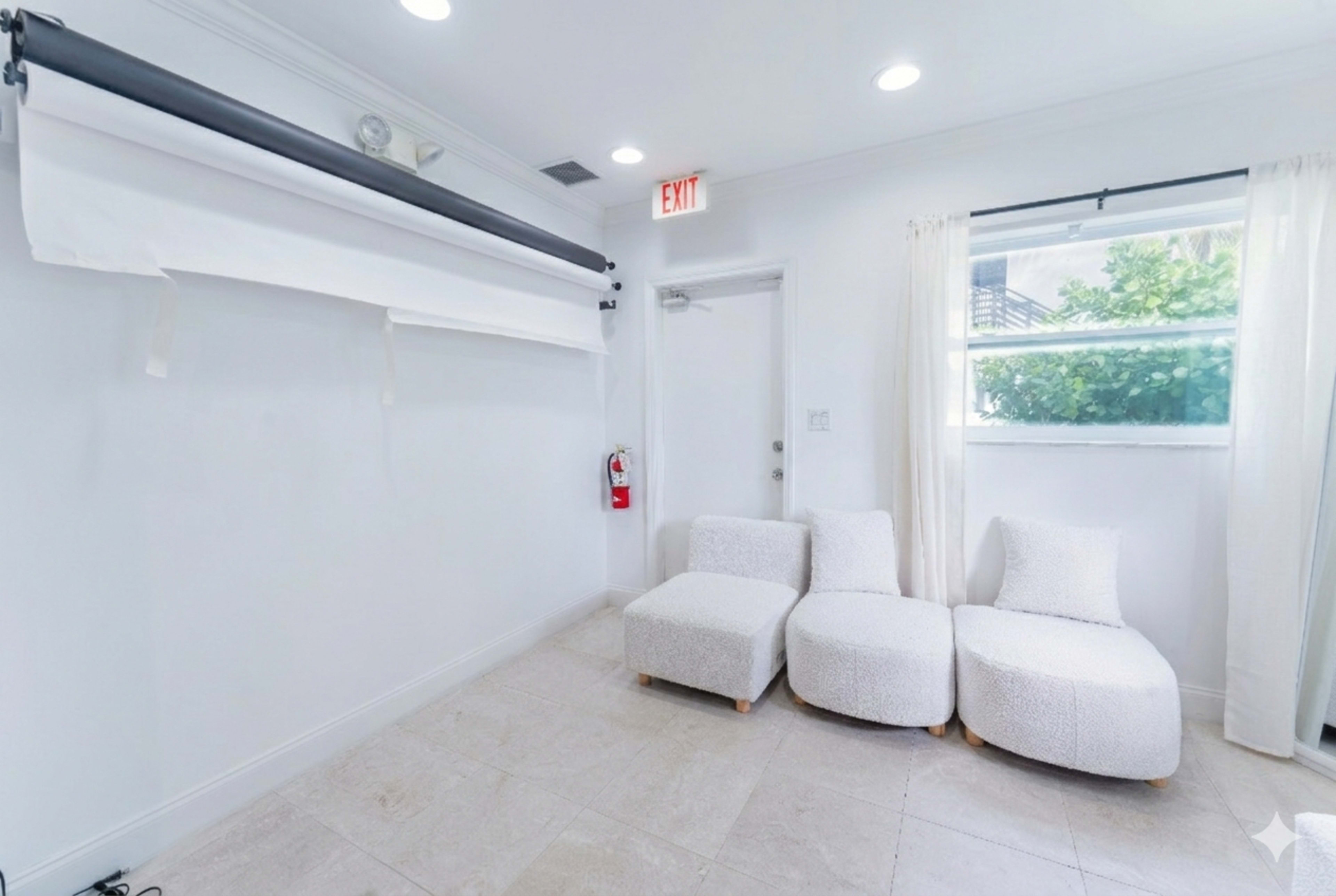 The image shows a bright, minimalist room featuring three white upholstered chairs and a wall with a roll-up projector screen.