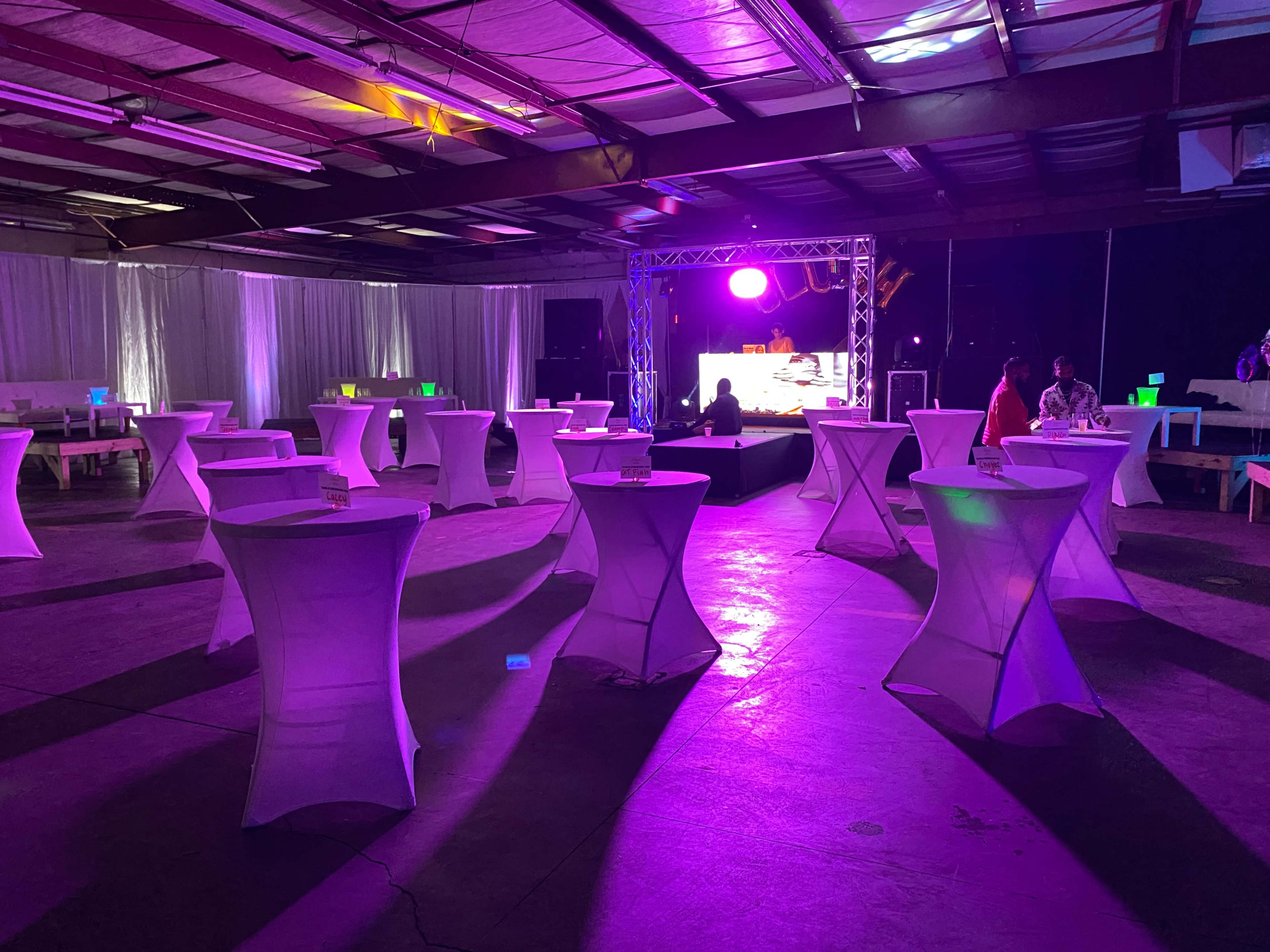 The image shows a spacious indoor venue set up for an event, featuring white cocktail tables and colorful lighting from a stage in the background.