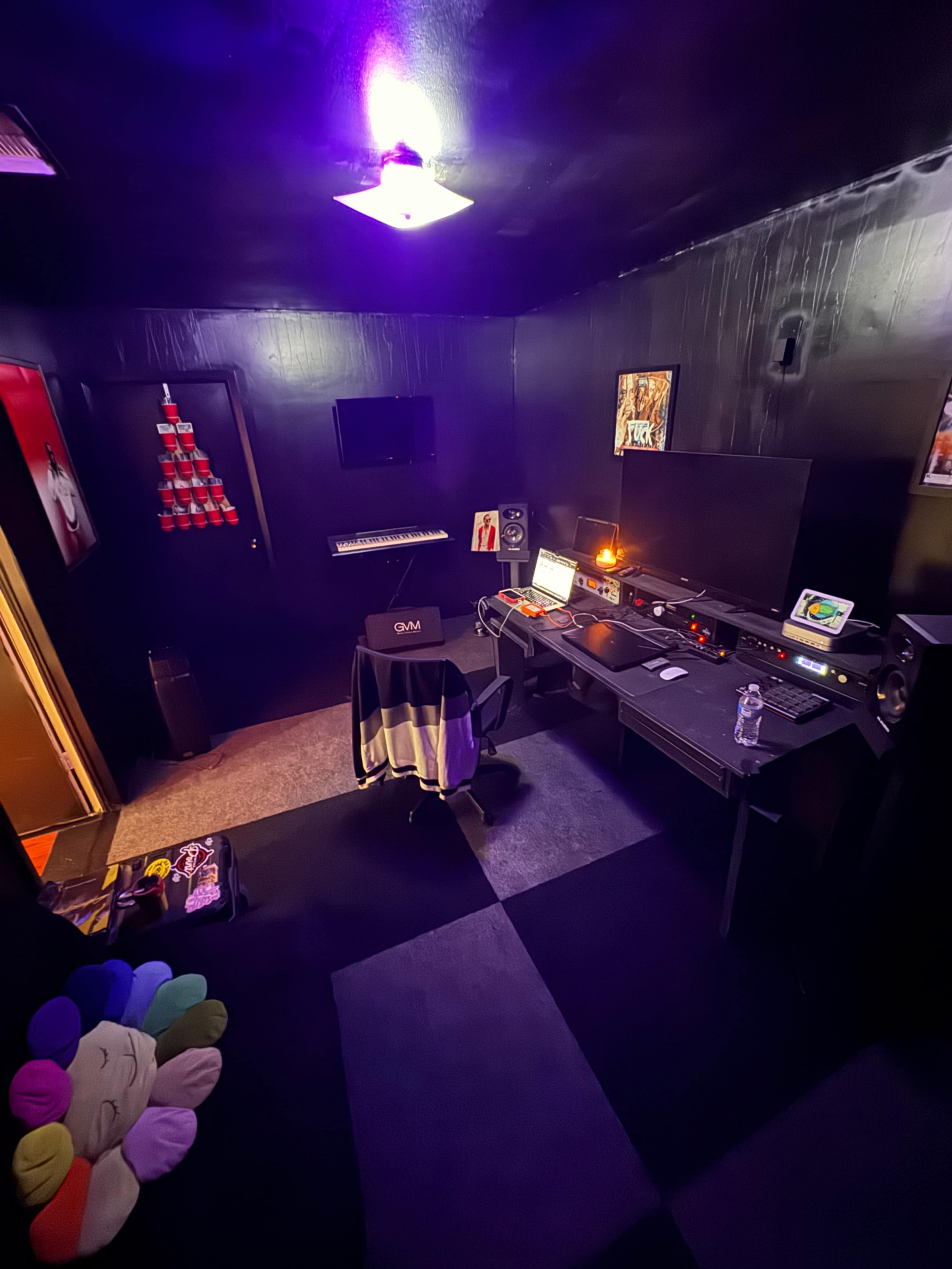 The image shows a music studio with dark walls, a computer desk equipped with sound monitors, a keyboard, and colorful lighting fixtures.