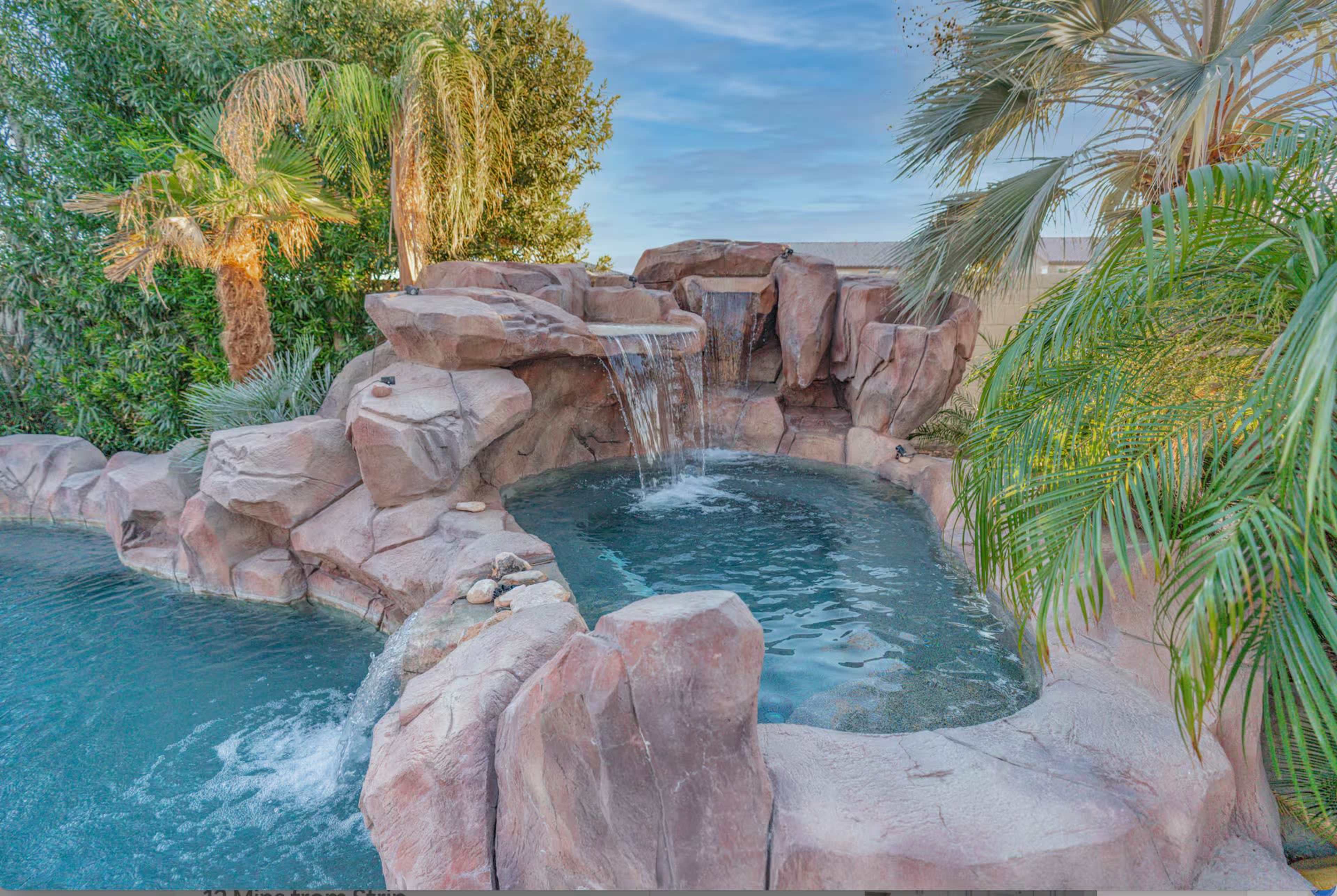 The image shows a rock formation with a waterfall cascading into a circular pool surrounded by palm trees.