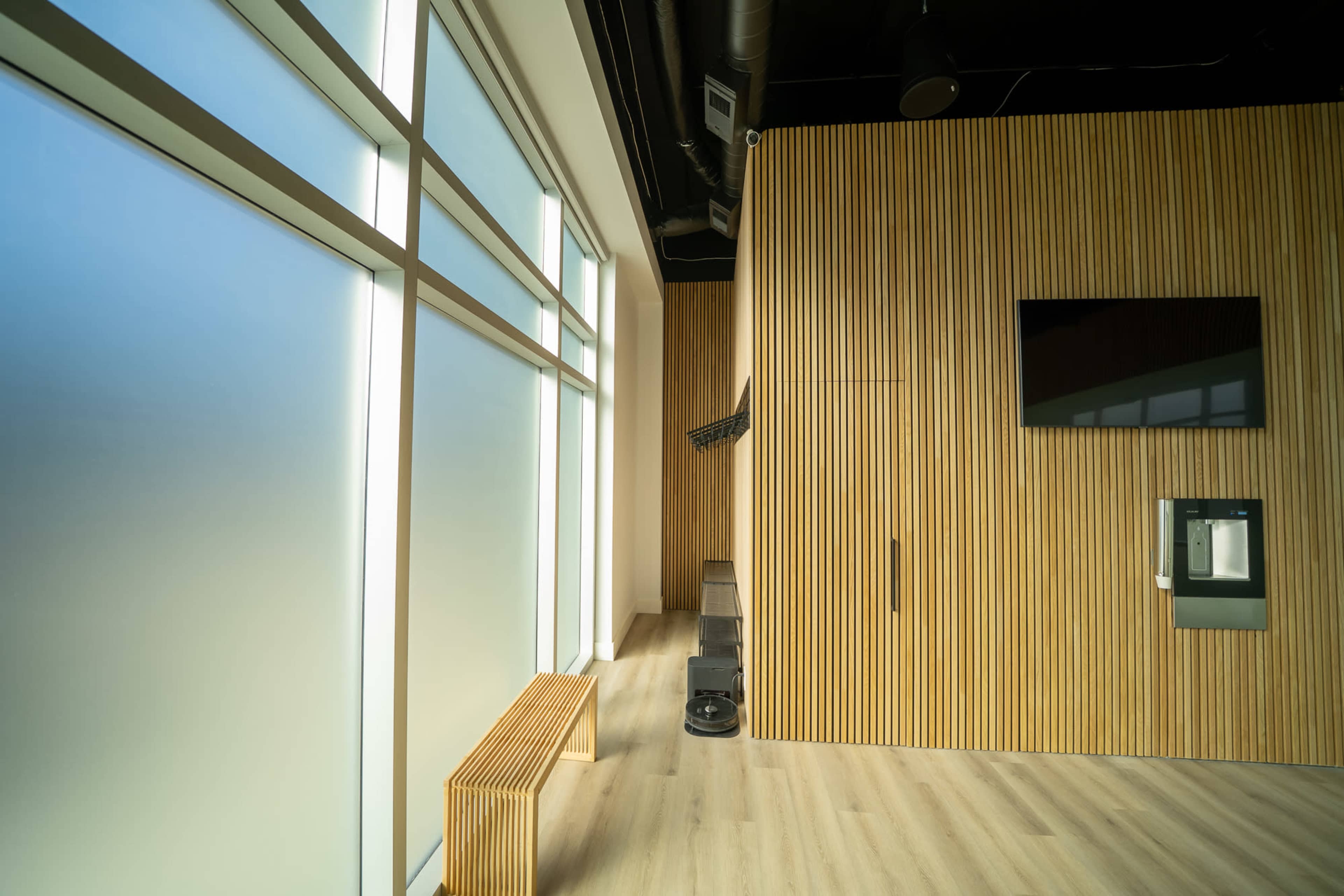 The image shows a minimalist interior space featuring wooden walls, large frosted windows, a bench, and a wall-mounted television.