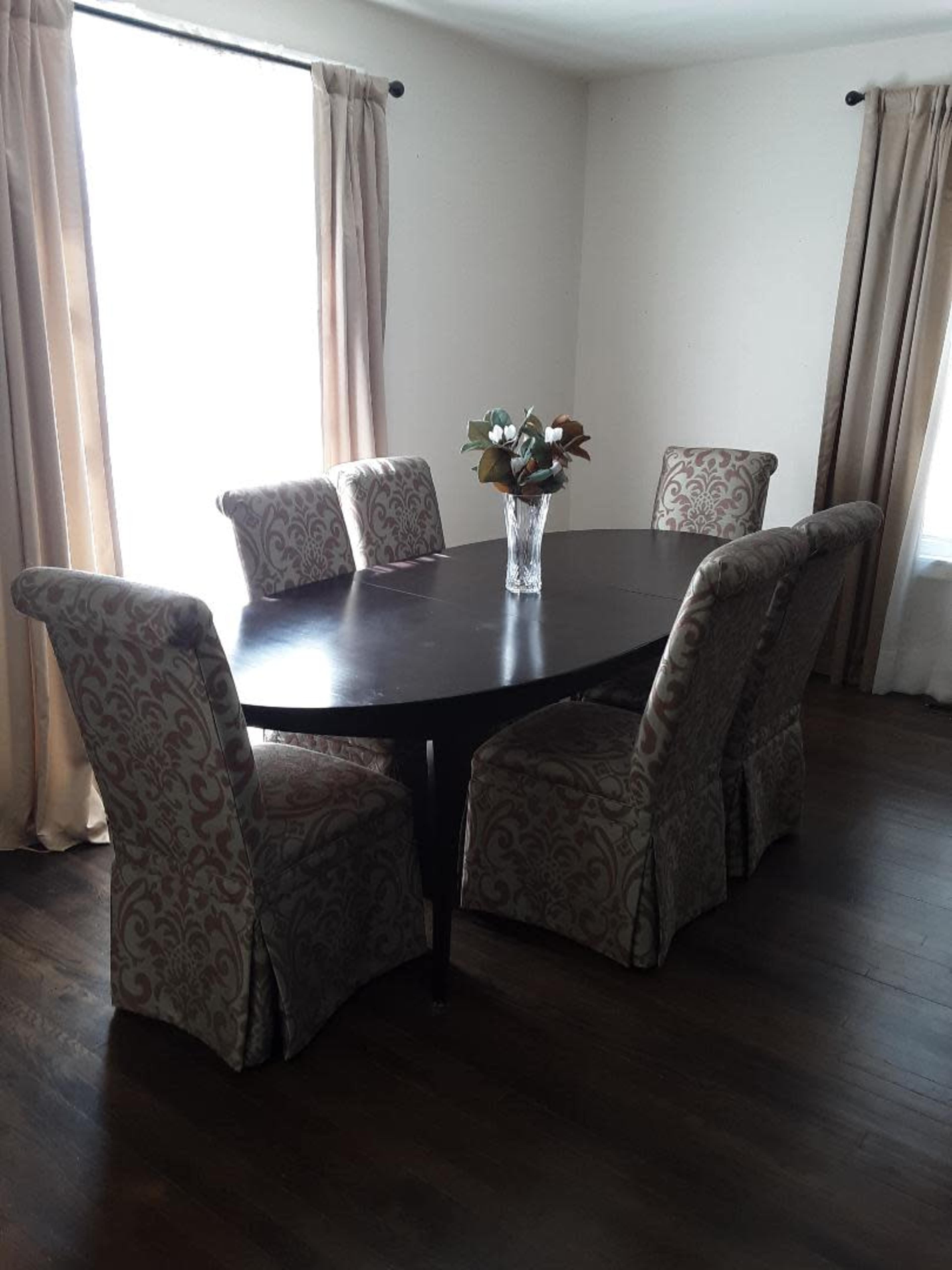 A wooden oval dining table is surrounded by six patterned upholstered chairs, with a vase of flowers in the center, set in a well-lit room with two windows.
