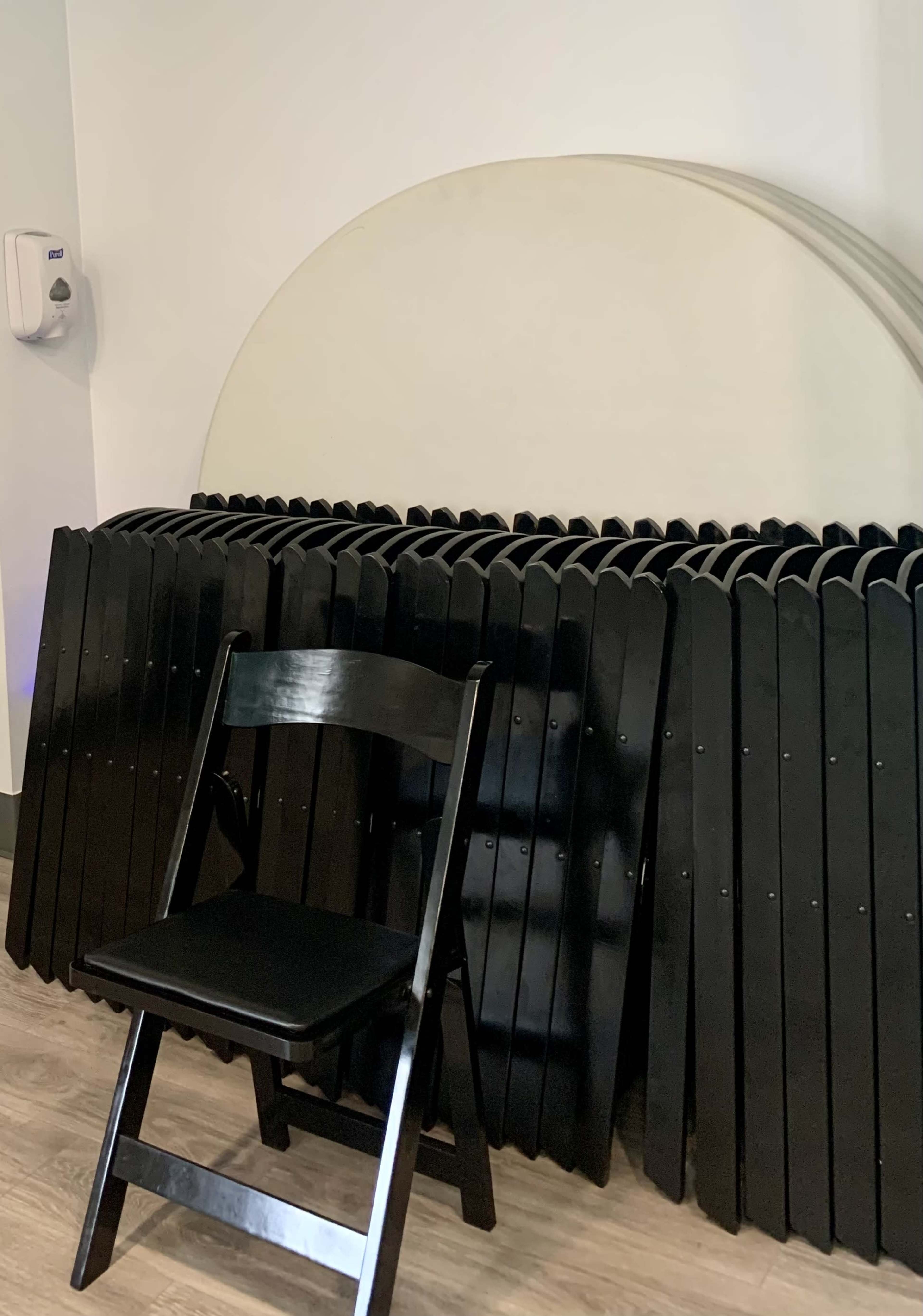 A black folding chair stands in front of a stacked row of black folding tables against a wall.