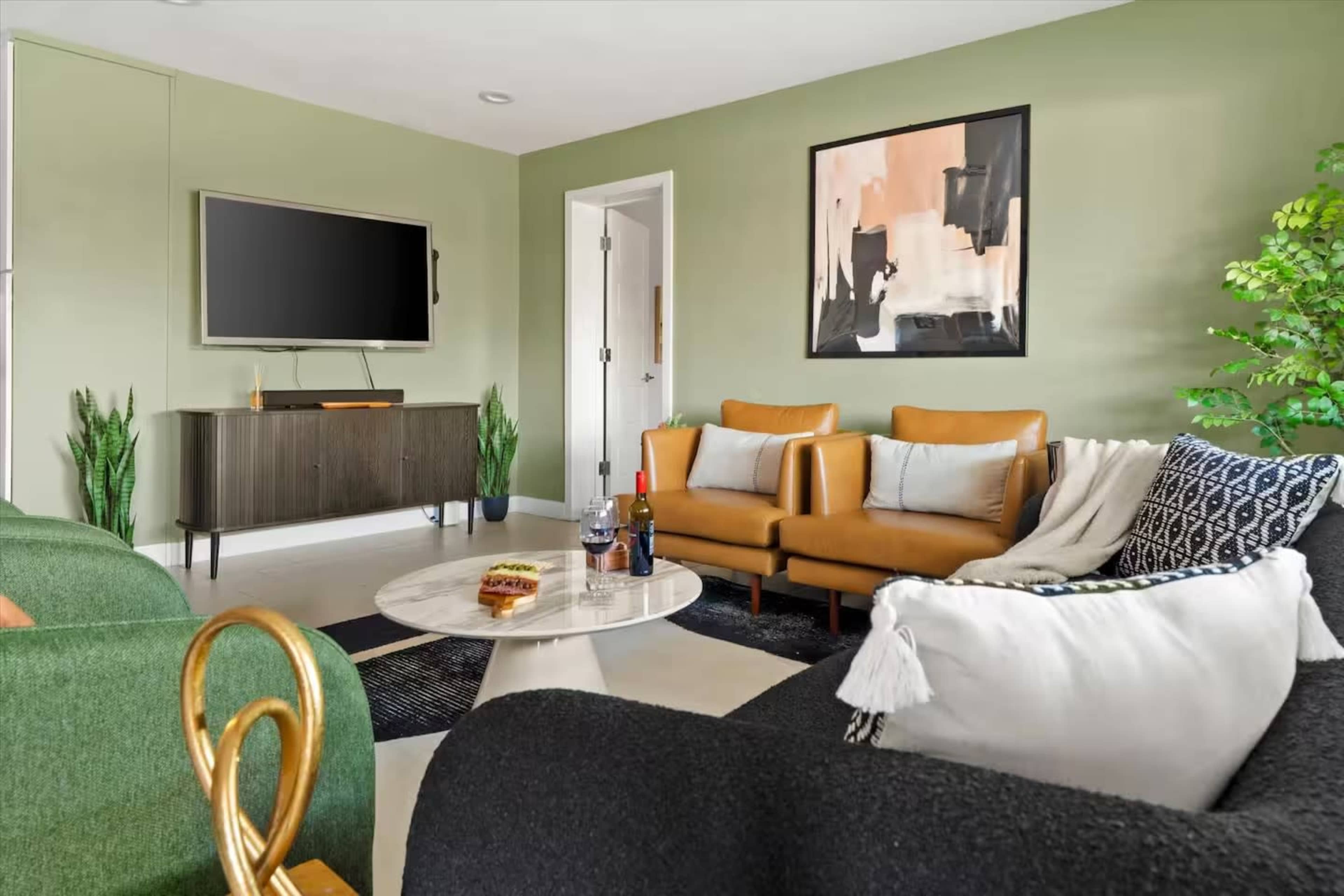 The image shows a modern living room with two brown leather chairs, a black sofa, a round marble coffee table, a television mounted on the wall, and green plants in the corners.