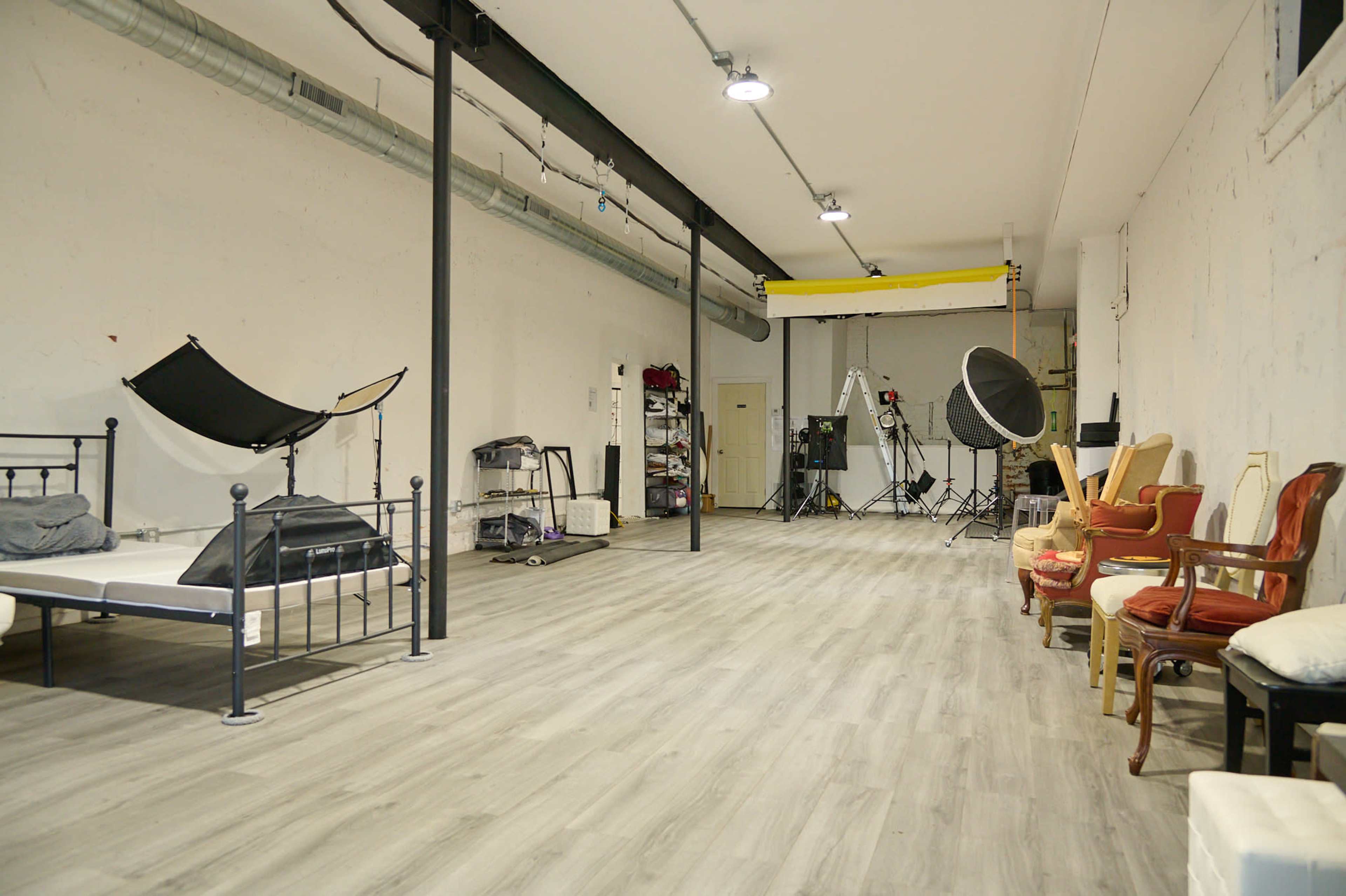 A large, empty studio space with a bed, various lighting equipment, and several chairs arranged along one side.