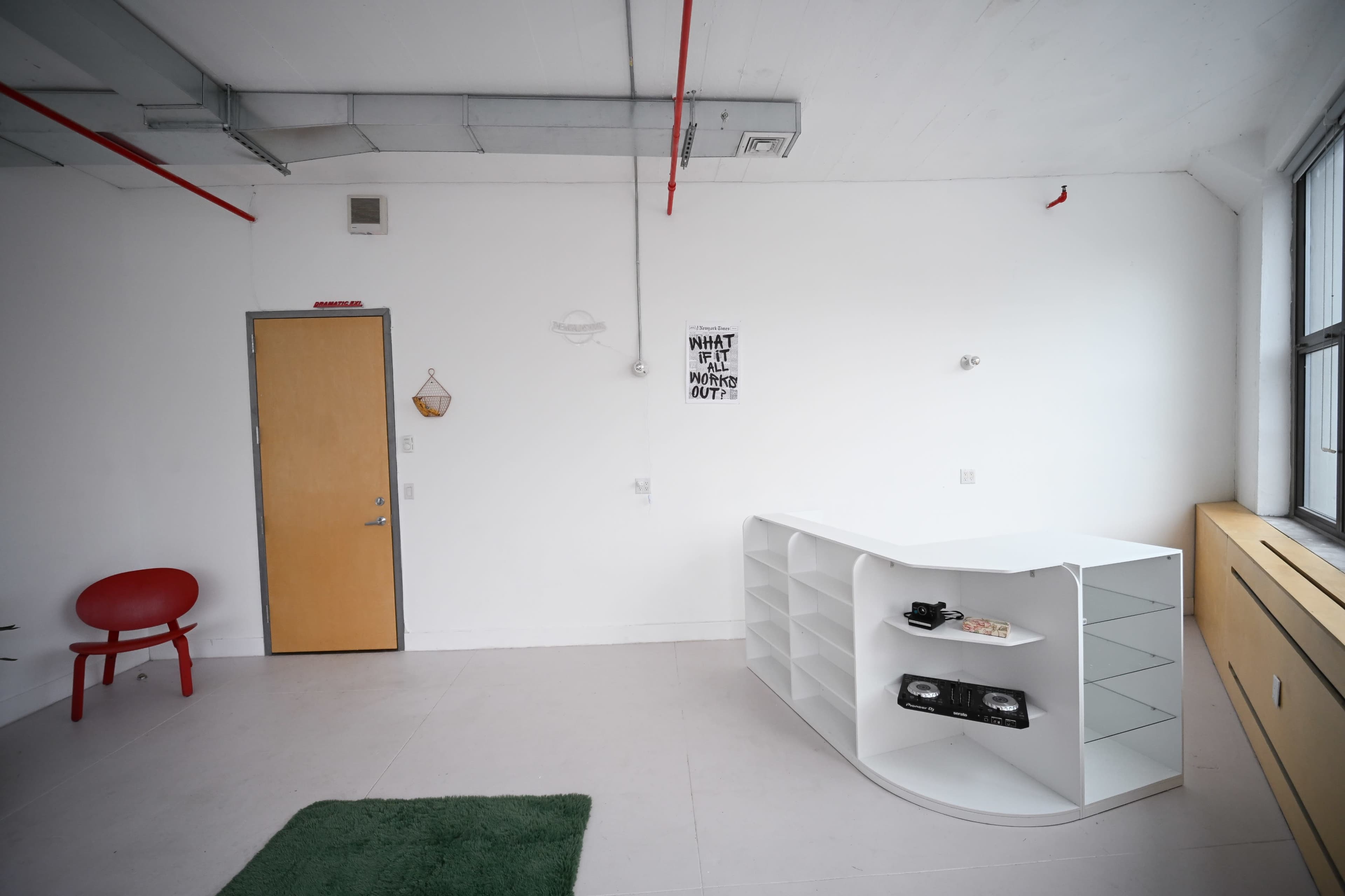 The image shows a minimalist studio space with a white wall, red piping, and a curved white shelving unit, alongside a red chair and a green rug on the floor.