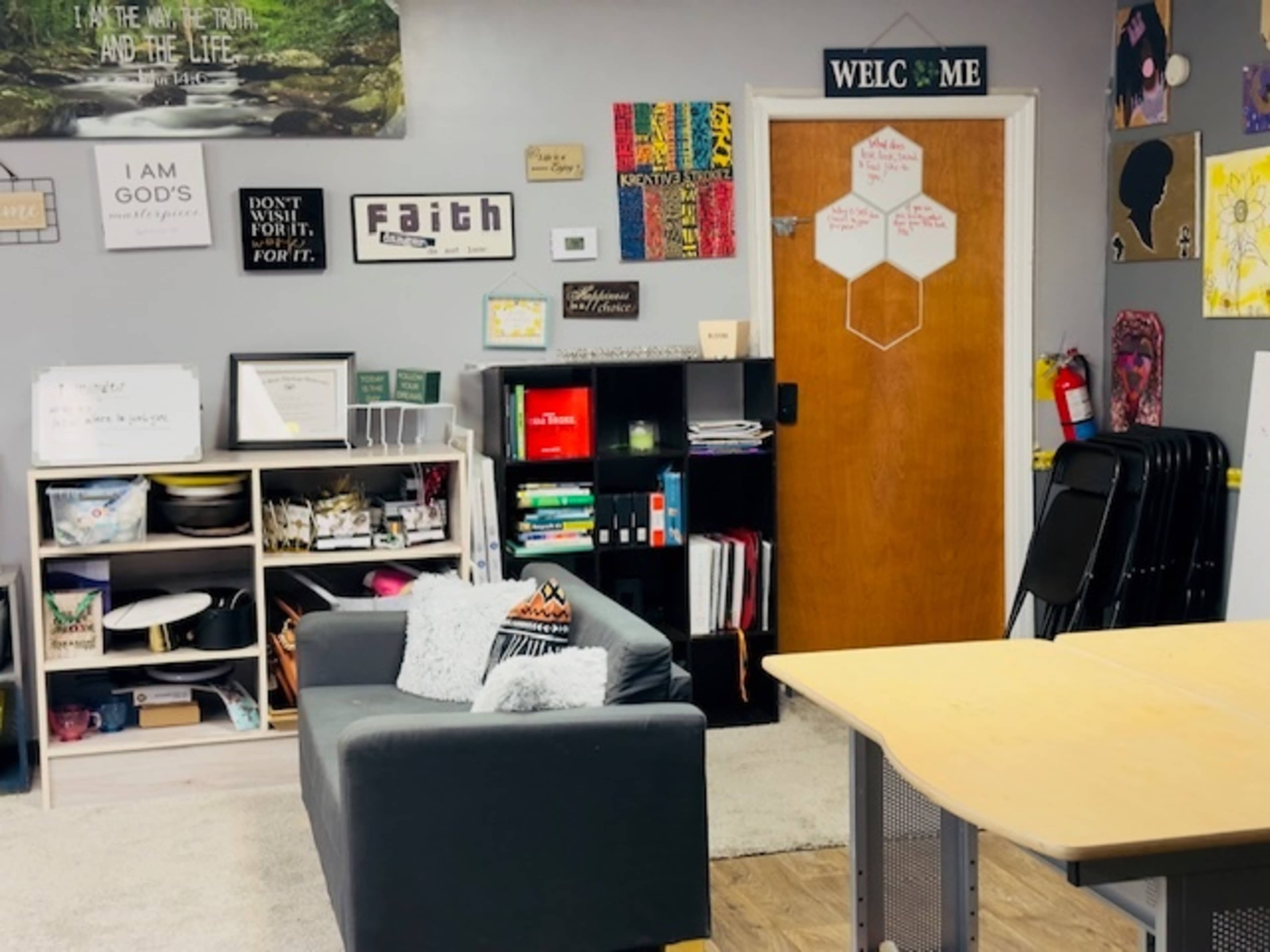 The image shows a classroom or office space with a black couch, a wooden table, shelves filled with books and supplies, and various wall decorations including posters and signs.