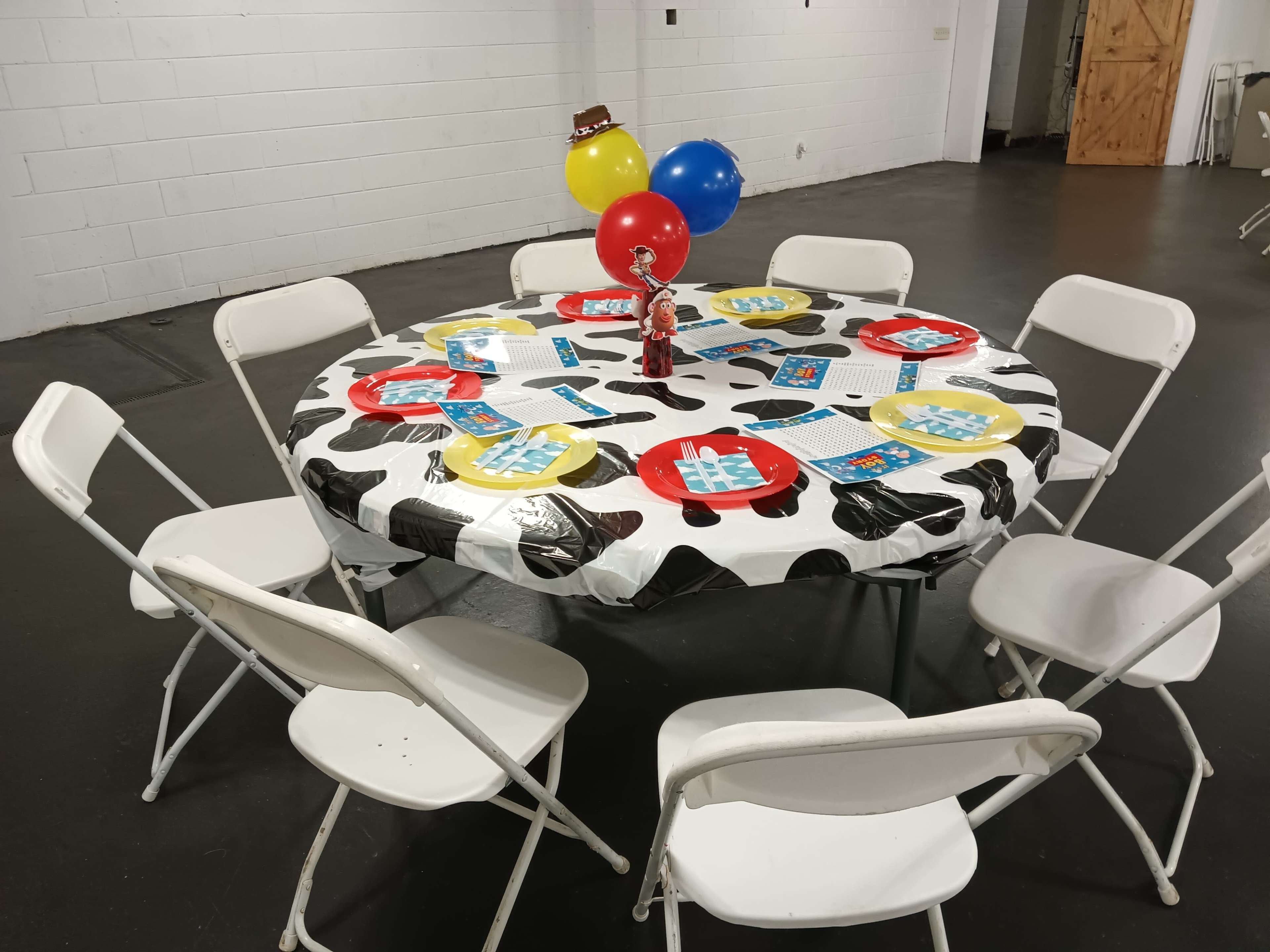 A round table covered with a cow-print tablecloth is set with red plates and balloons, surrounded by white folding chairs in a plain room.