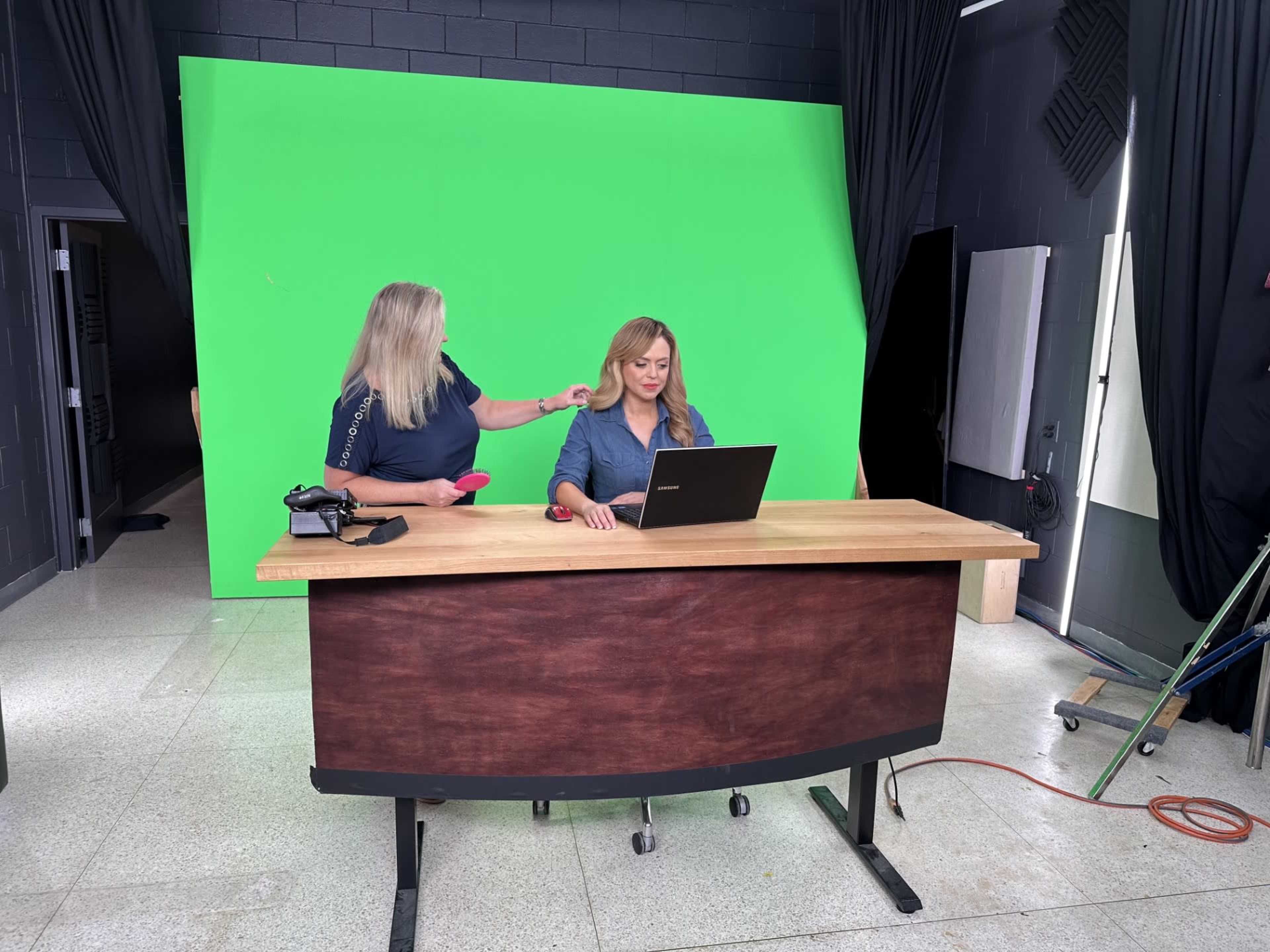 A woman sits at a desk with a laptop while another woman adjusts her hair in front of a green screen backdrop.