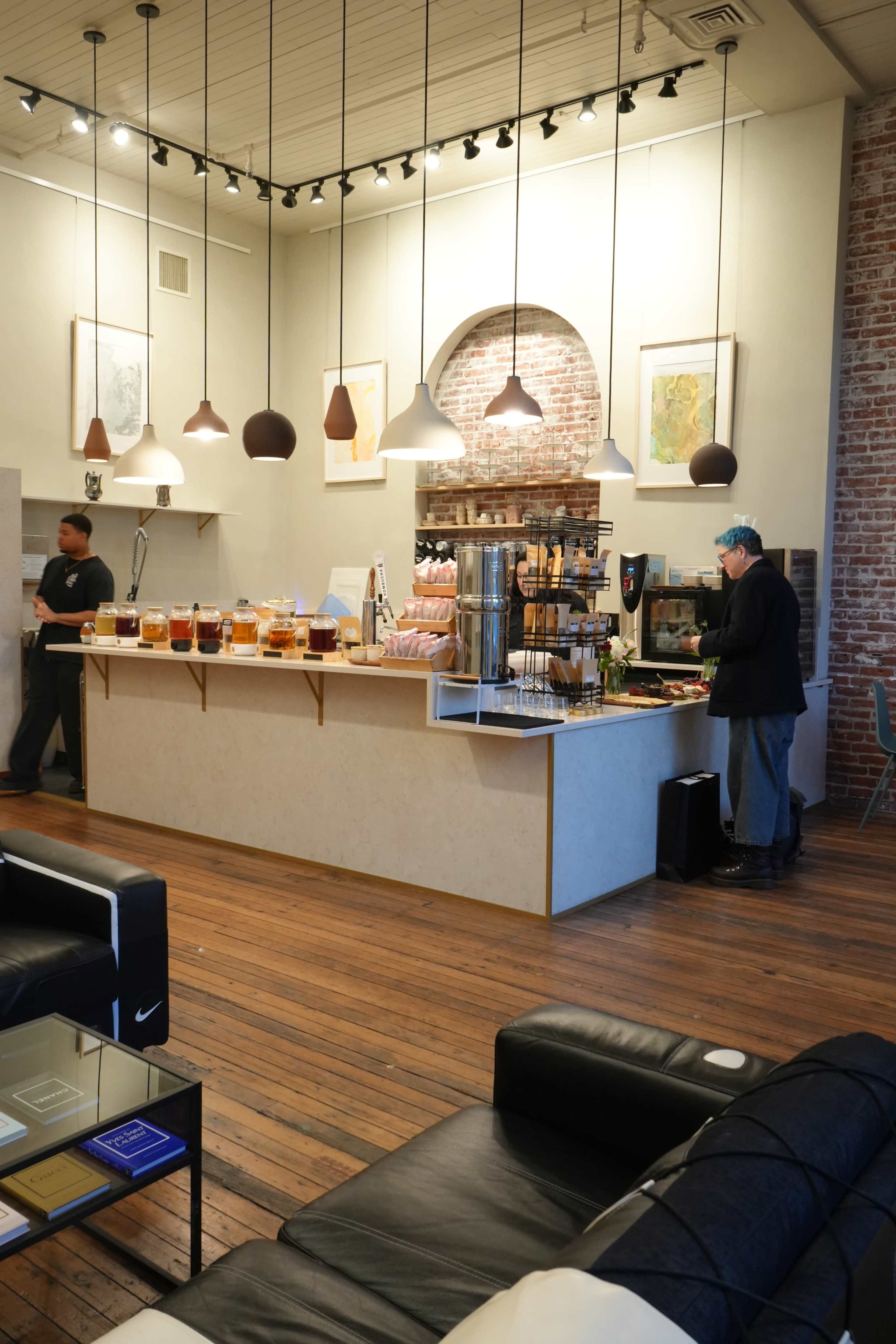 A spacious café features a counter with various beverage dispensers and pastries, while two employees attend to customers in a bright, well-lit environment.