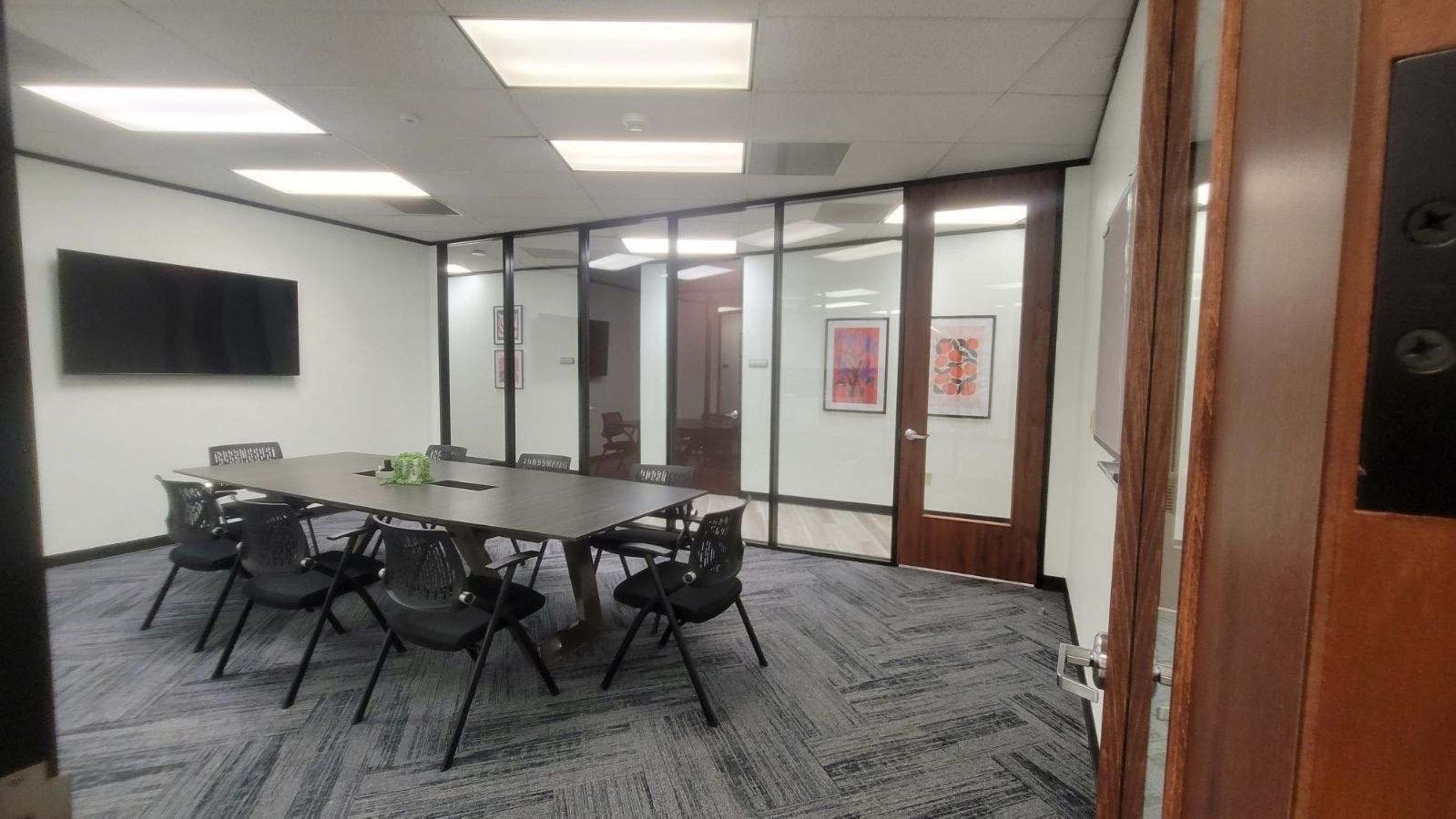 The image shows a modern conference room featuring a long table surrounded by black chairs, with glass walls and a television mounted on one side.