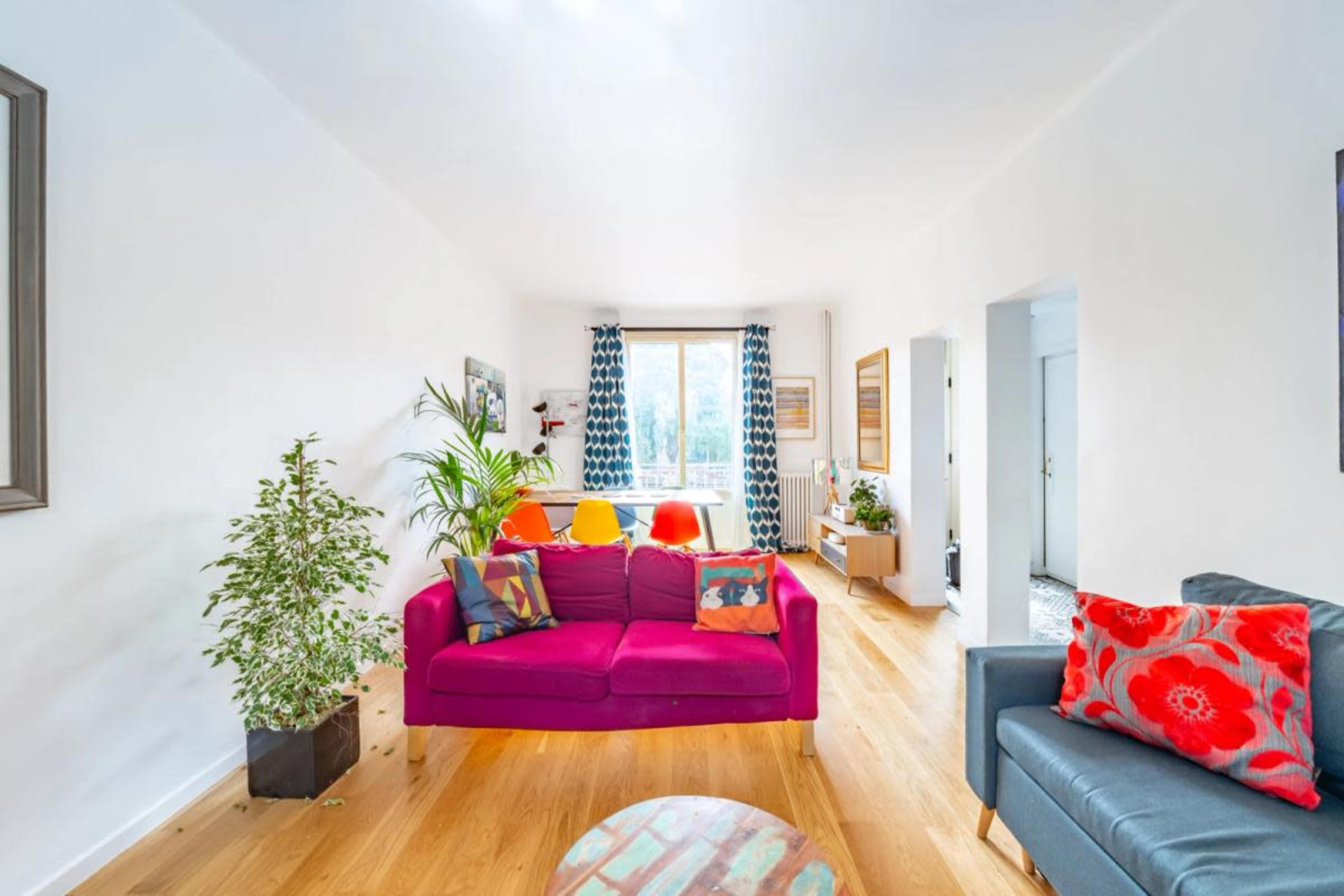 A bright living room features a purple sofa, a gray armchair, and large windows dressed with patterned curtains, surrounded by indoor plants and wooden flooring.