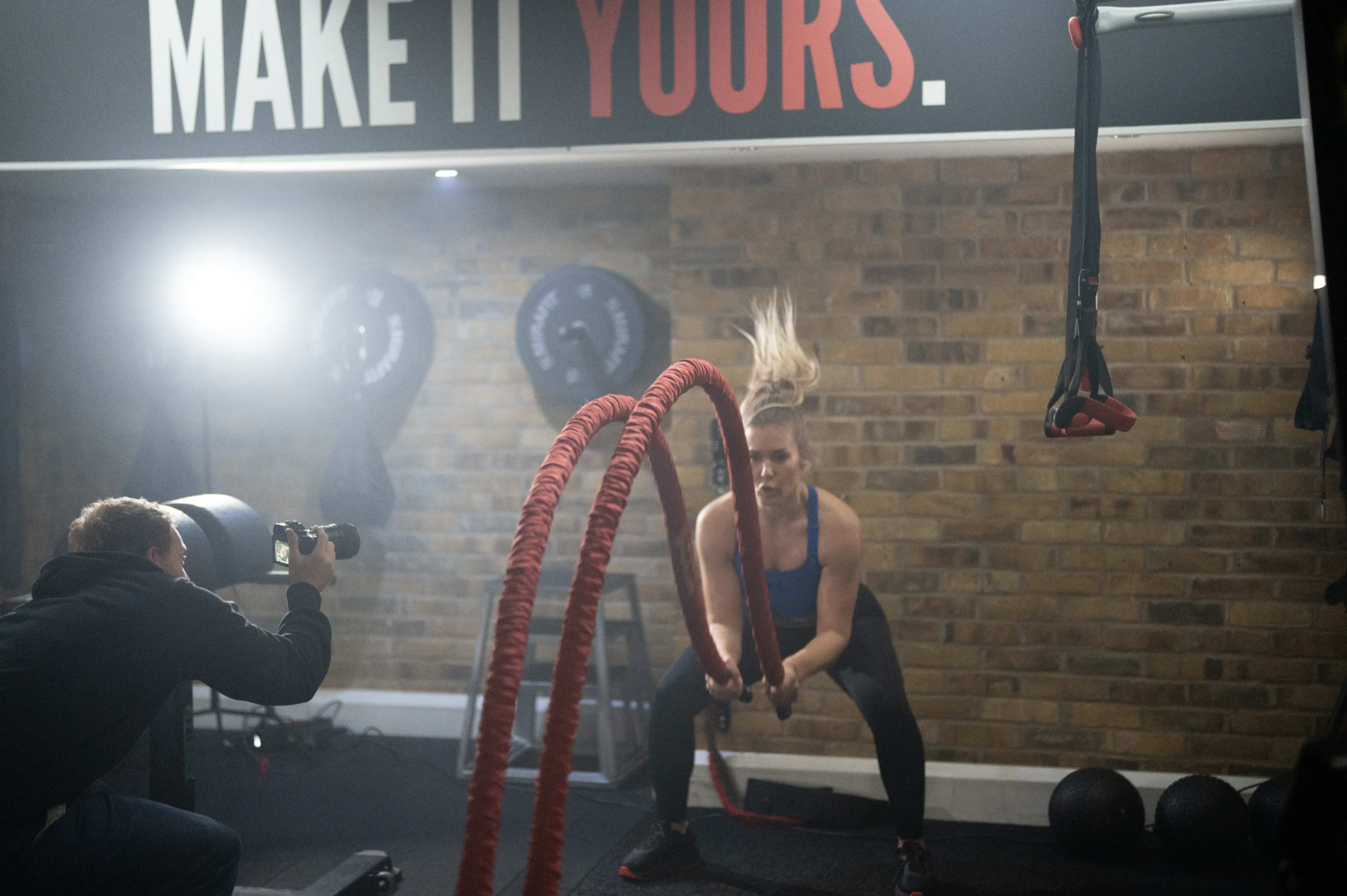 A woman performs battle rope exercises in a gym while a photographer captures the action.
