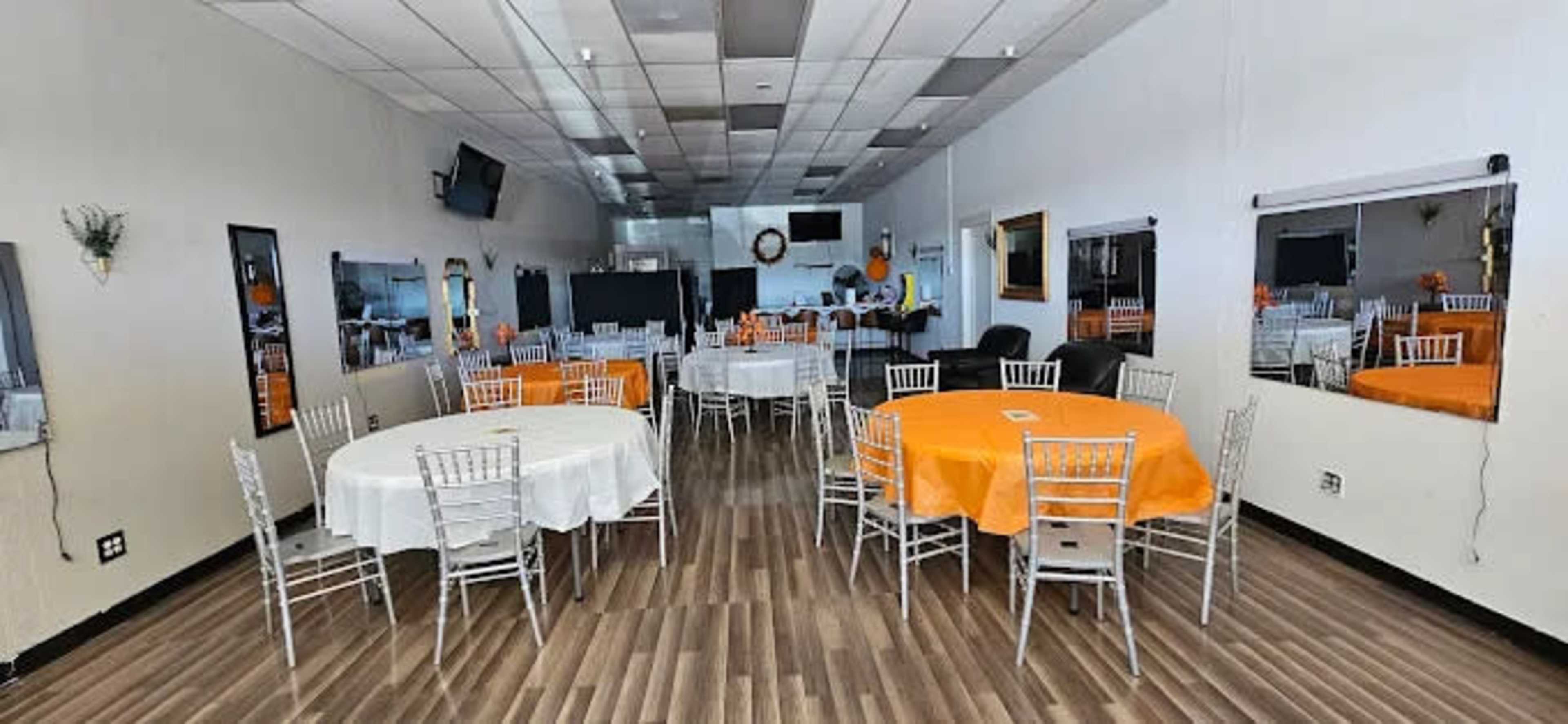 The image shows a spacious, well-lit event room furnished with round tables covered in orange and white tablecloths, surrounded by silver chiavari chairs.