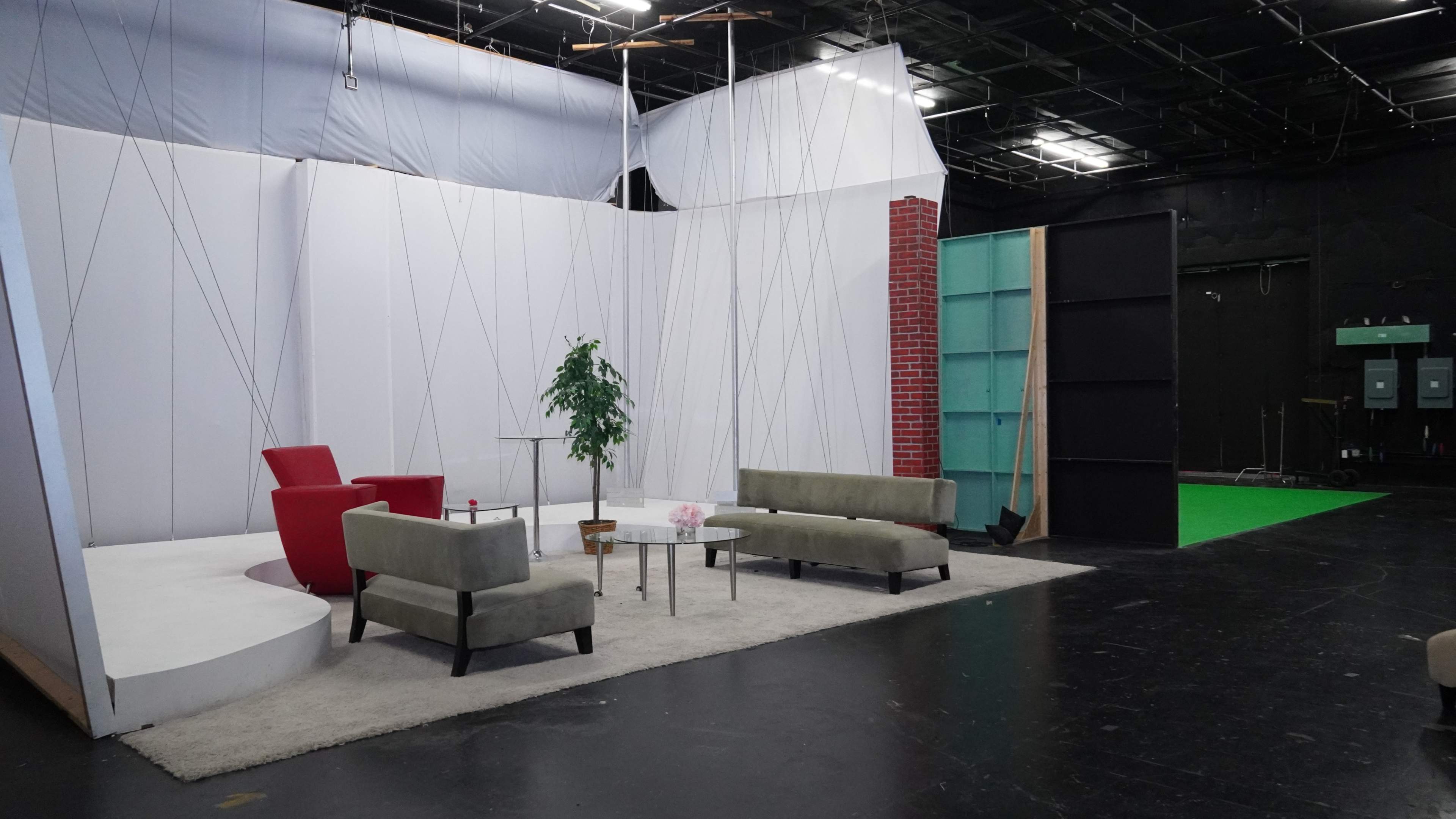 The image shows a modern television studio set featuring a circular white stage with a red chair, green couches, a small table, and a backdrop with a brick wall and a green screen area.