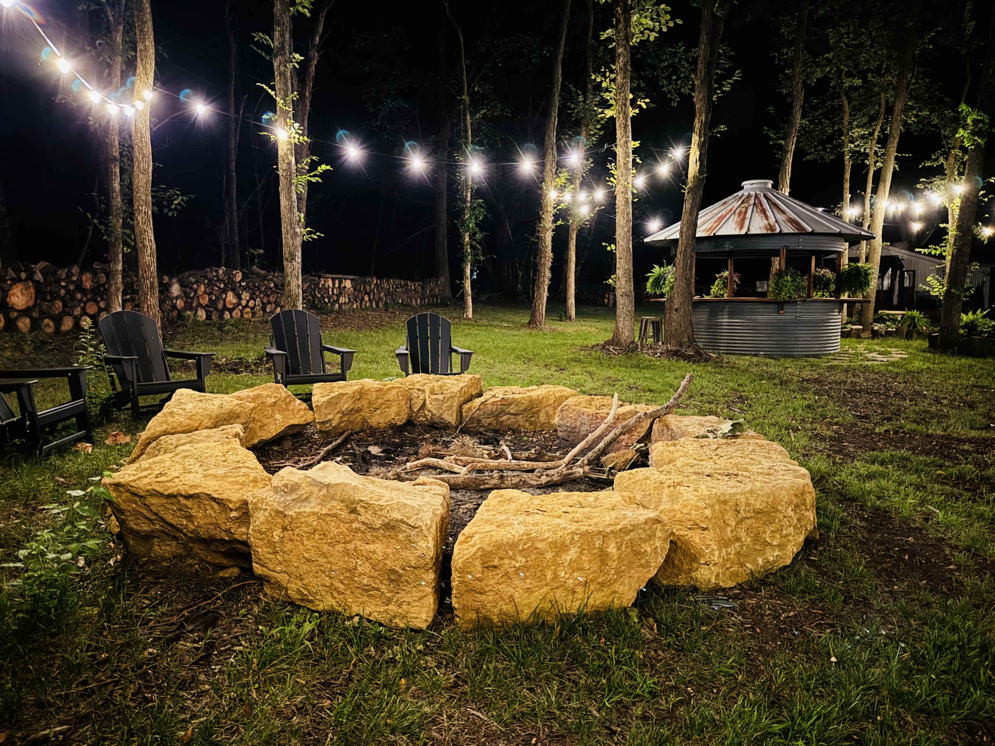 A stone fire pit surrounded by wooden chairs is positioned in a wooded area adorned with string lights, with a circular structure in the background.