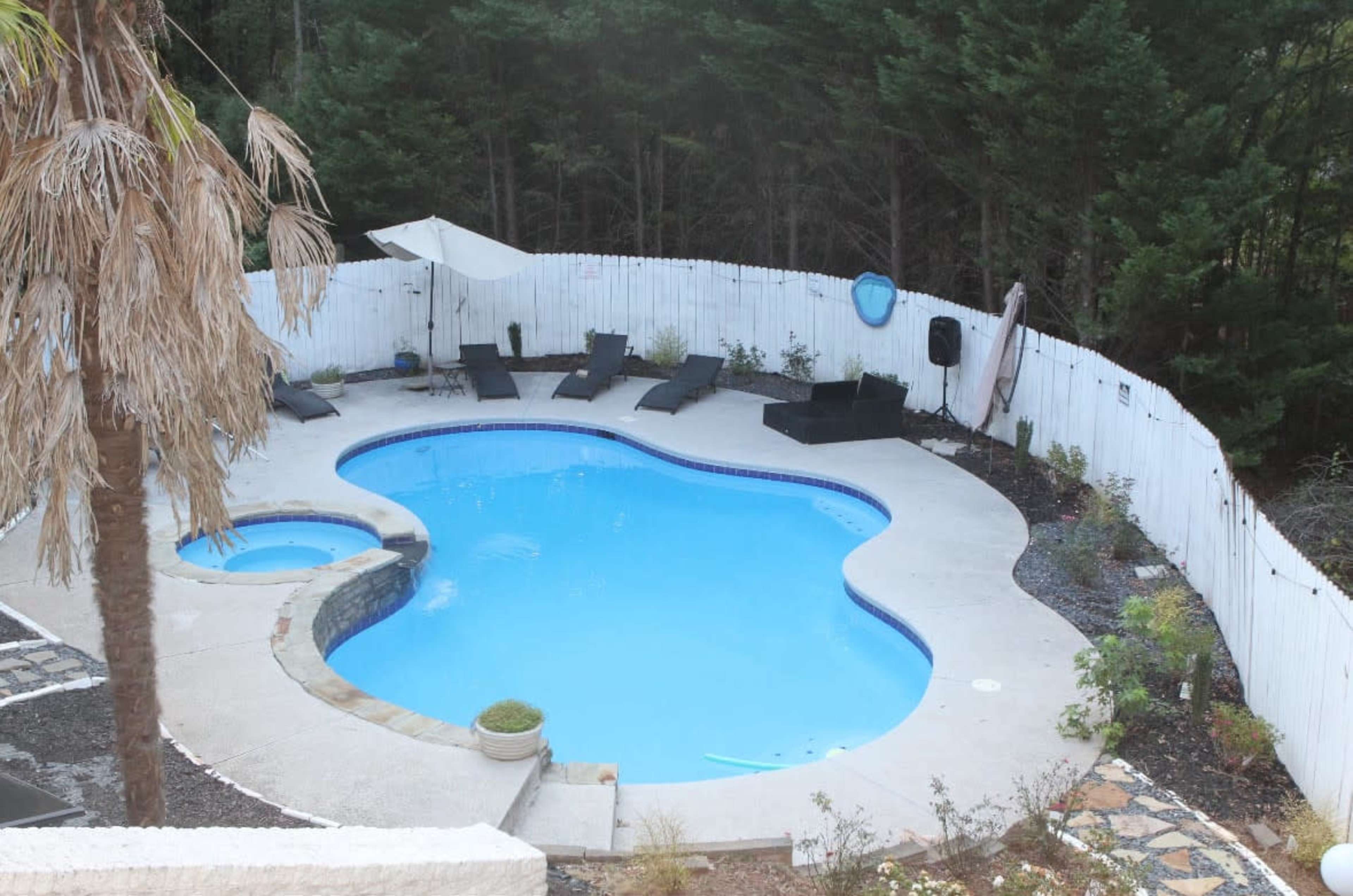 The image shows a swimming pool in a landscaped area surrounded by a white fence and greenery.