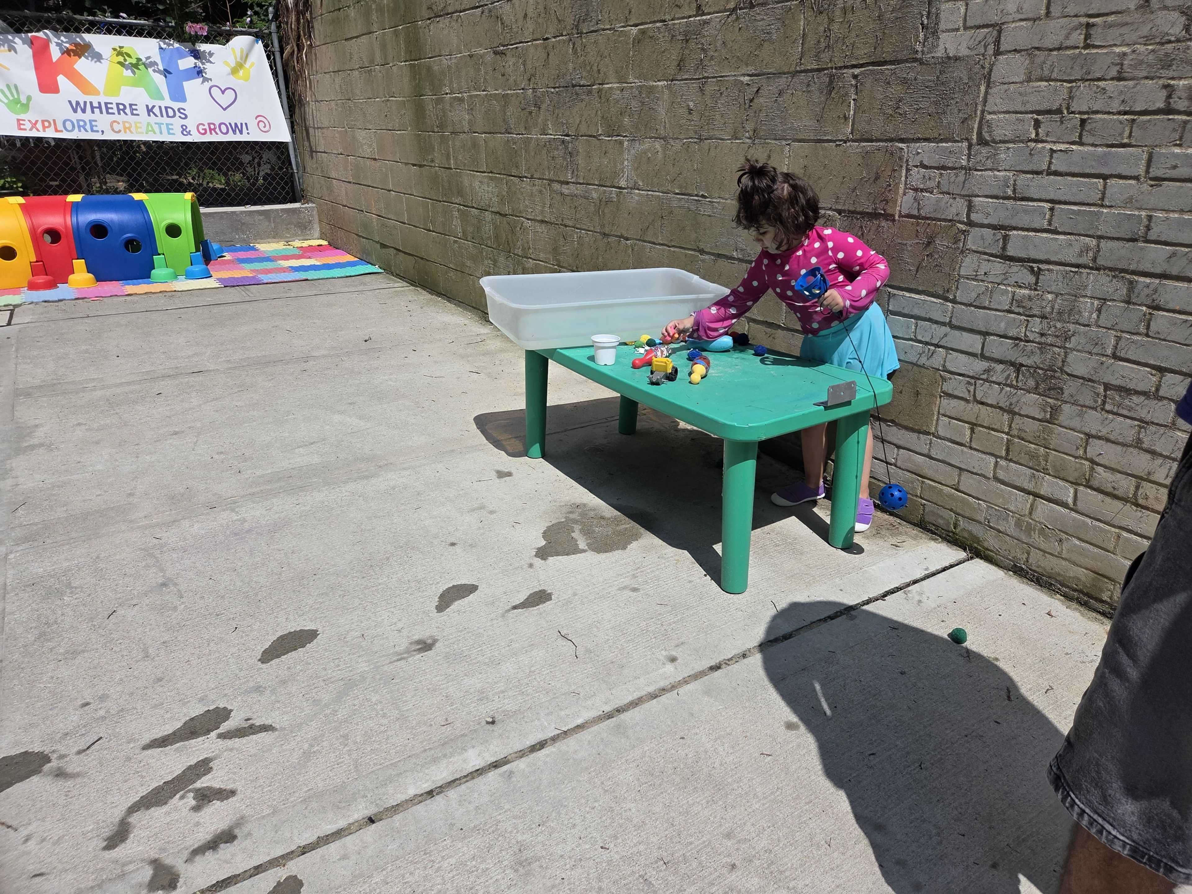 A child in a blue skirt plays with colorful toys at a green table next to a water tub on a concrete surface.