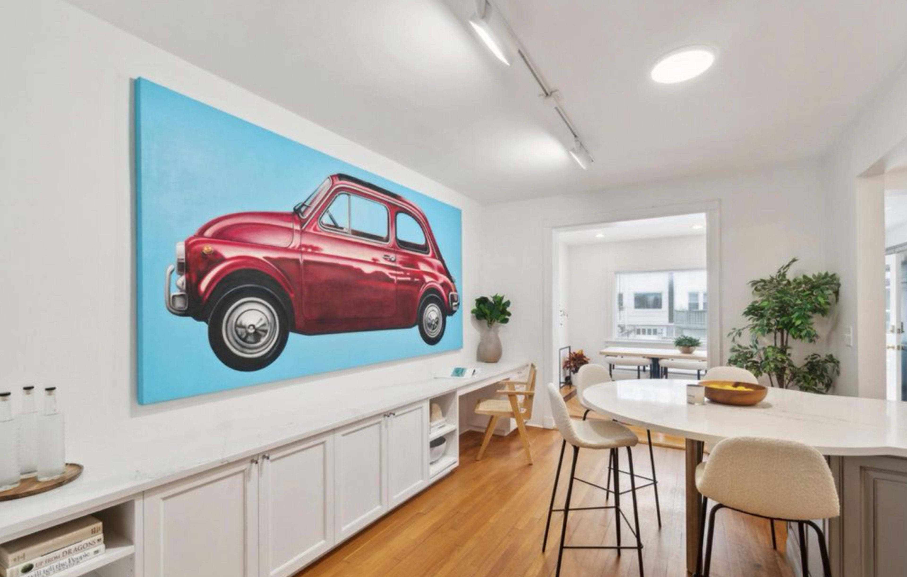 A bright and modern open-concept kitchen features a large painting of a red vintage car on a blue background, along with sleek furniture and plants.