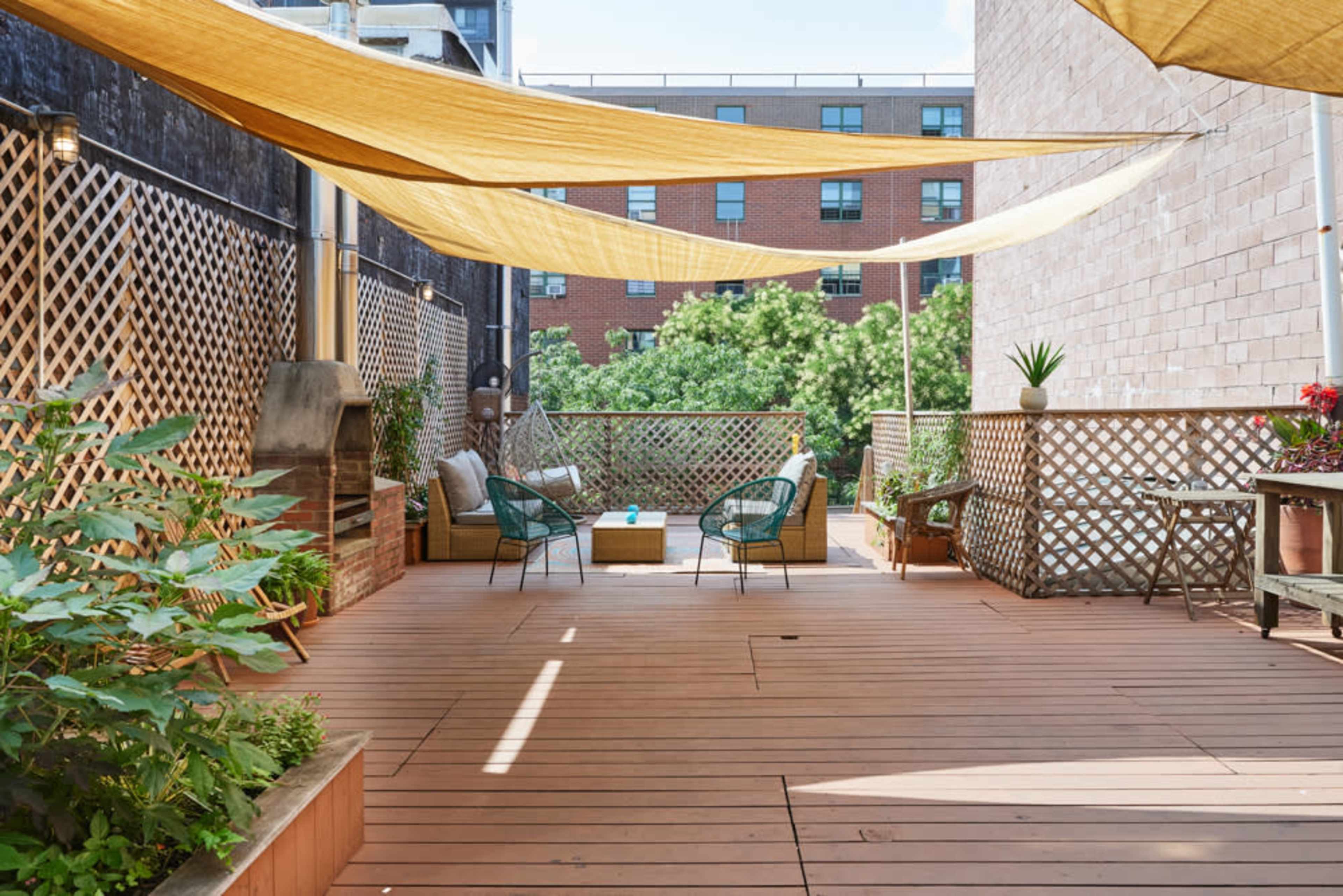 A wooden deck is shaded by yellow fabric canopies, featuring two chairs, a low table, and greenery along the edges.