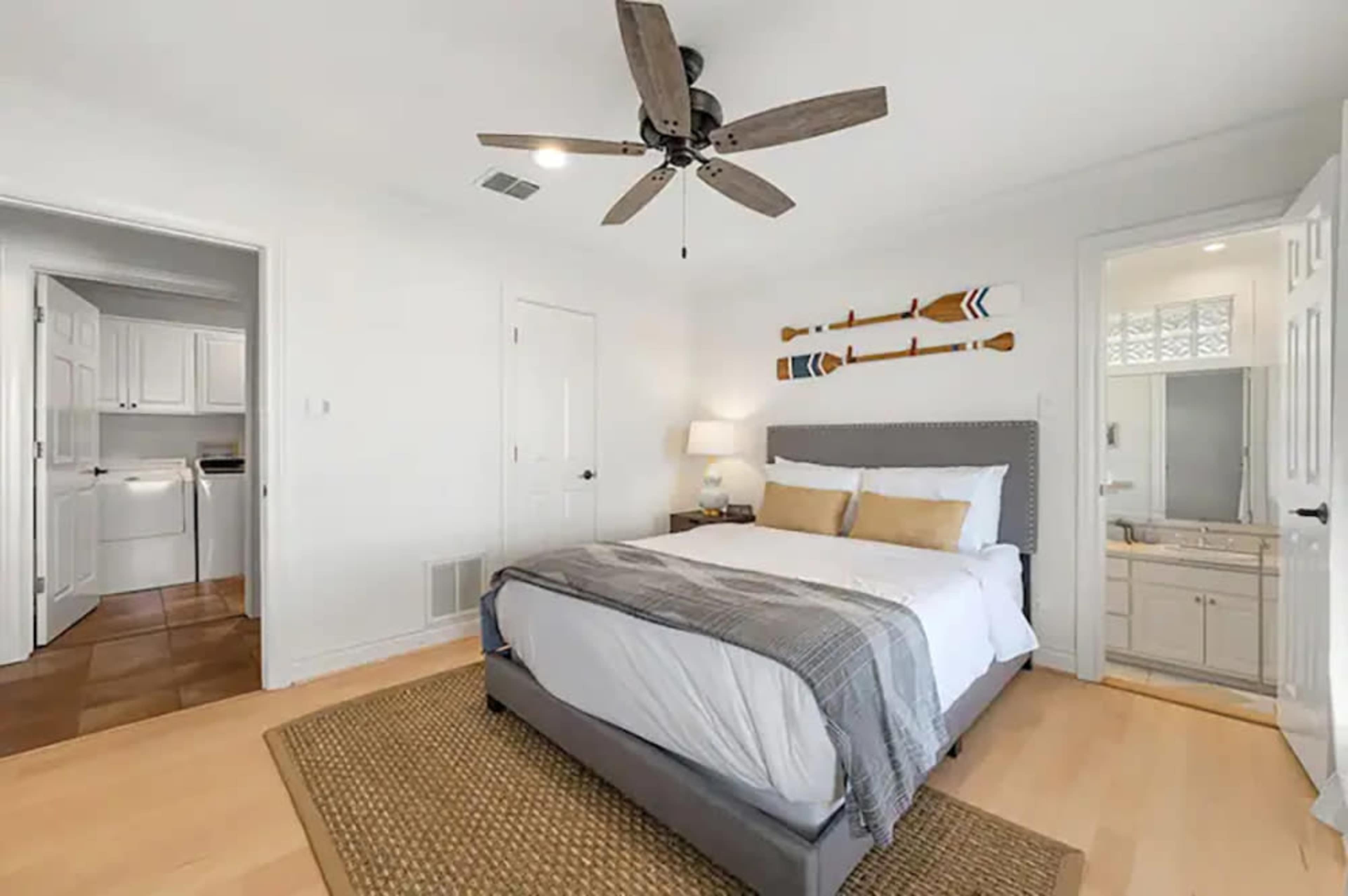A bright, minimalist bedroom featuring a queen-sized bed with gray and white bedding, a ceiling fan, and access to a bathroom and laundry area through two doors.