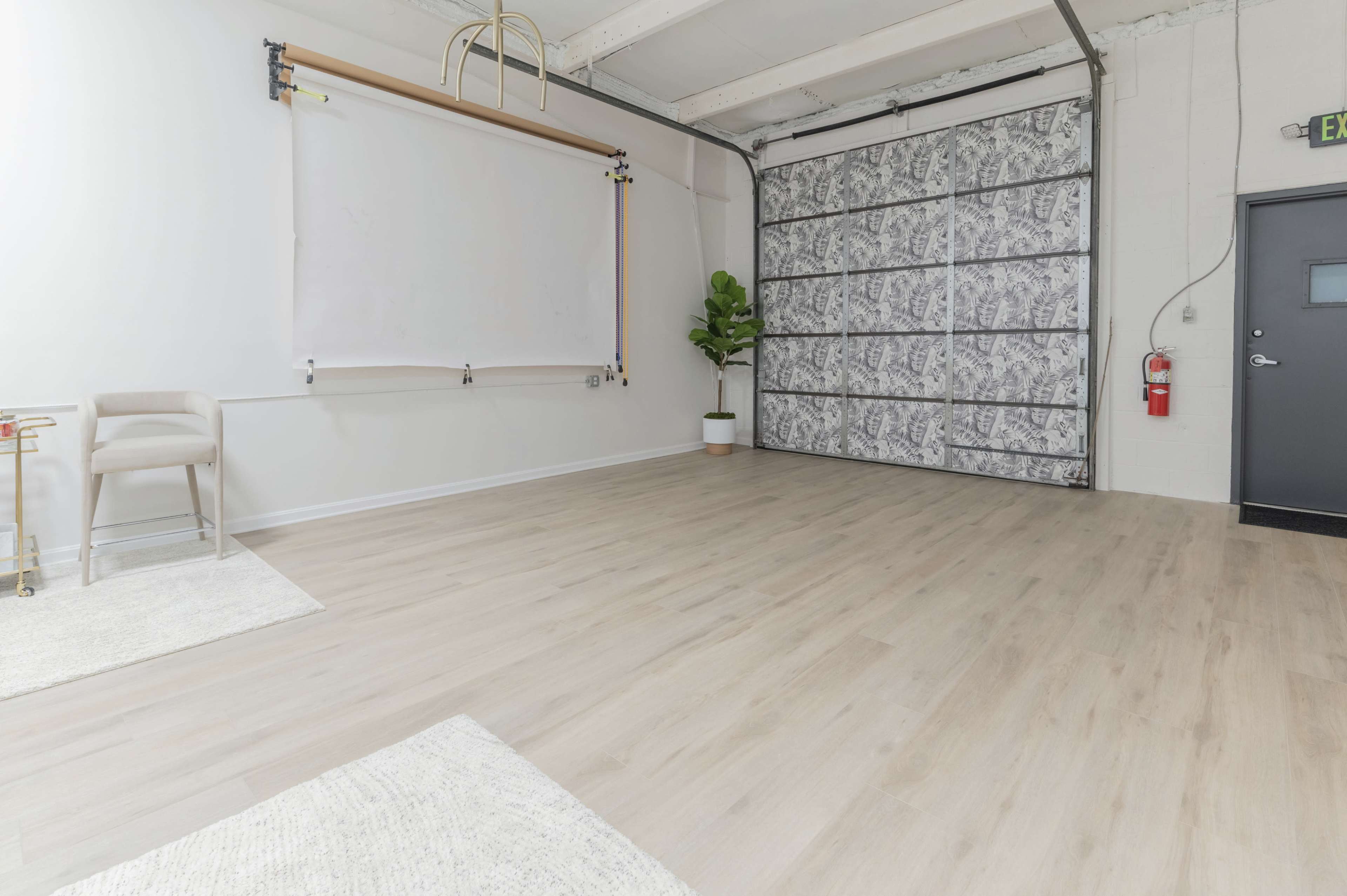 A spacious studio with light-colored wooden flooring, a white backdrop on a wall, a potted plant in the corner, and a large gray garage door.