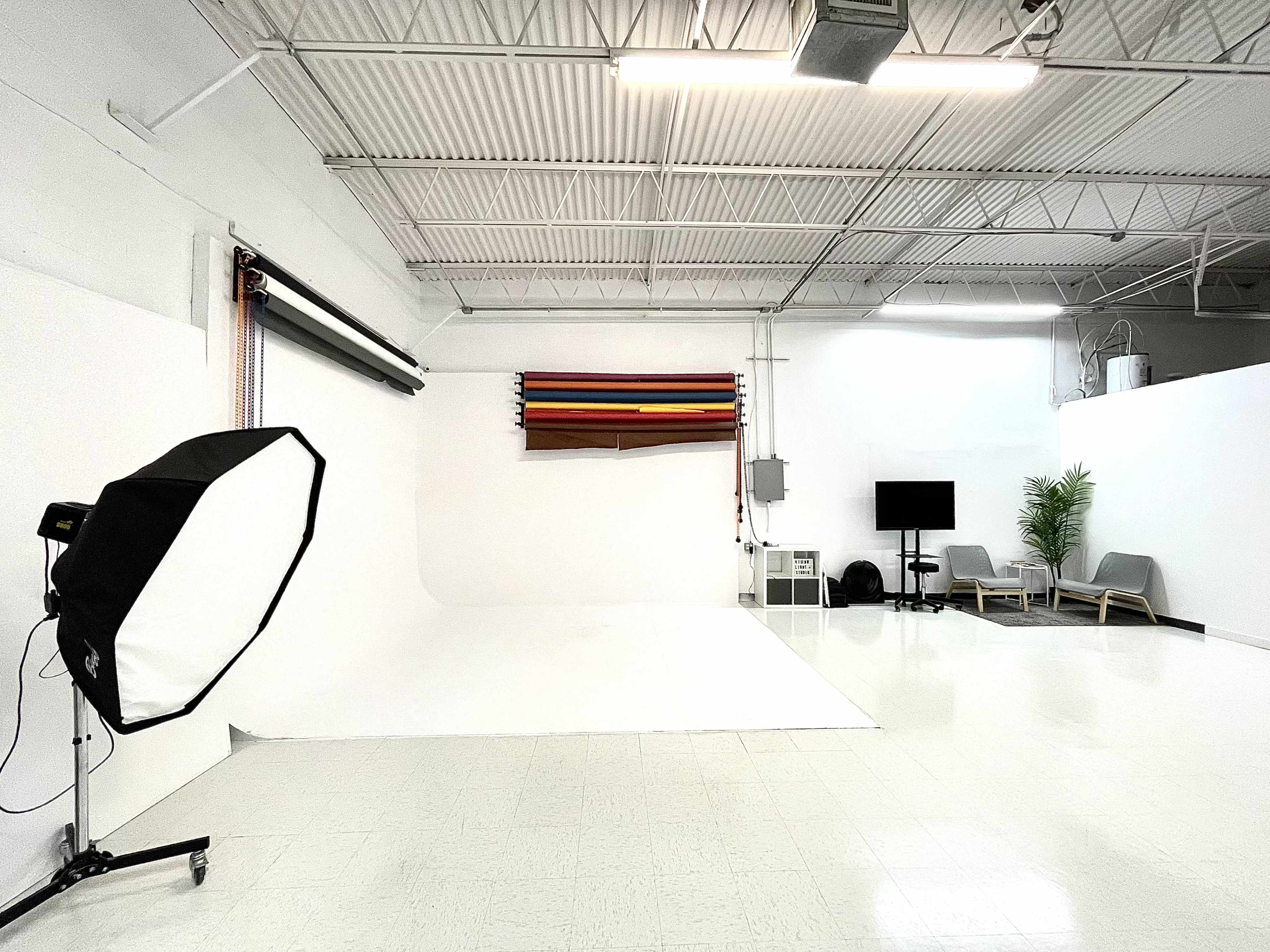 A spacious photography studio features a white backdrop, a lighting setup, and minimal furniture, including a television and a potted plant.