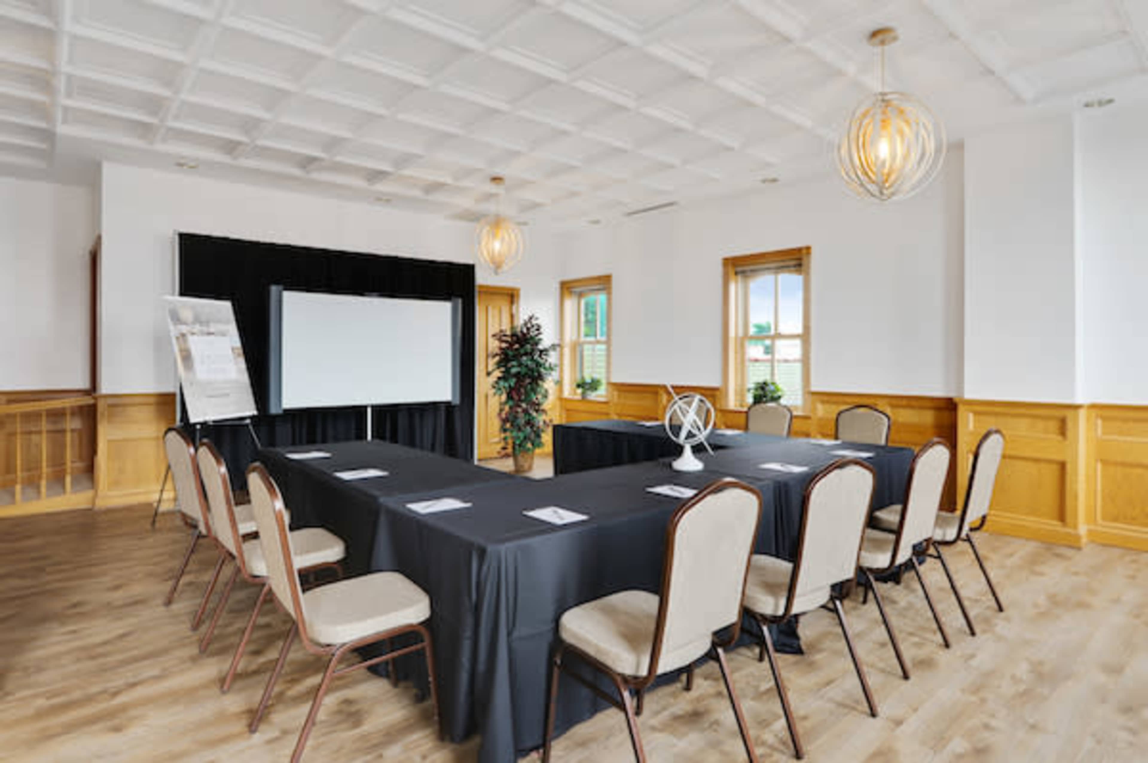 A conference room features a long black table surrounded by beige chairs, with a large screen at one end and windows allowing natural light.
