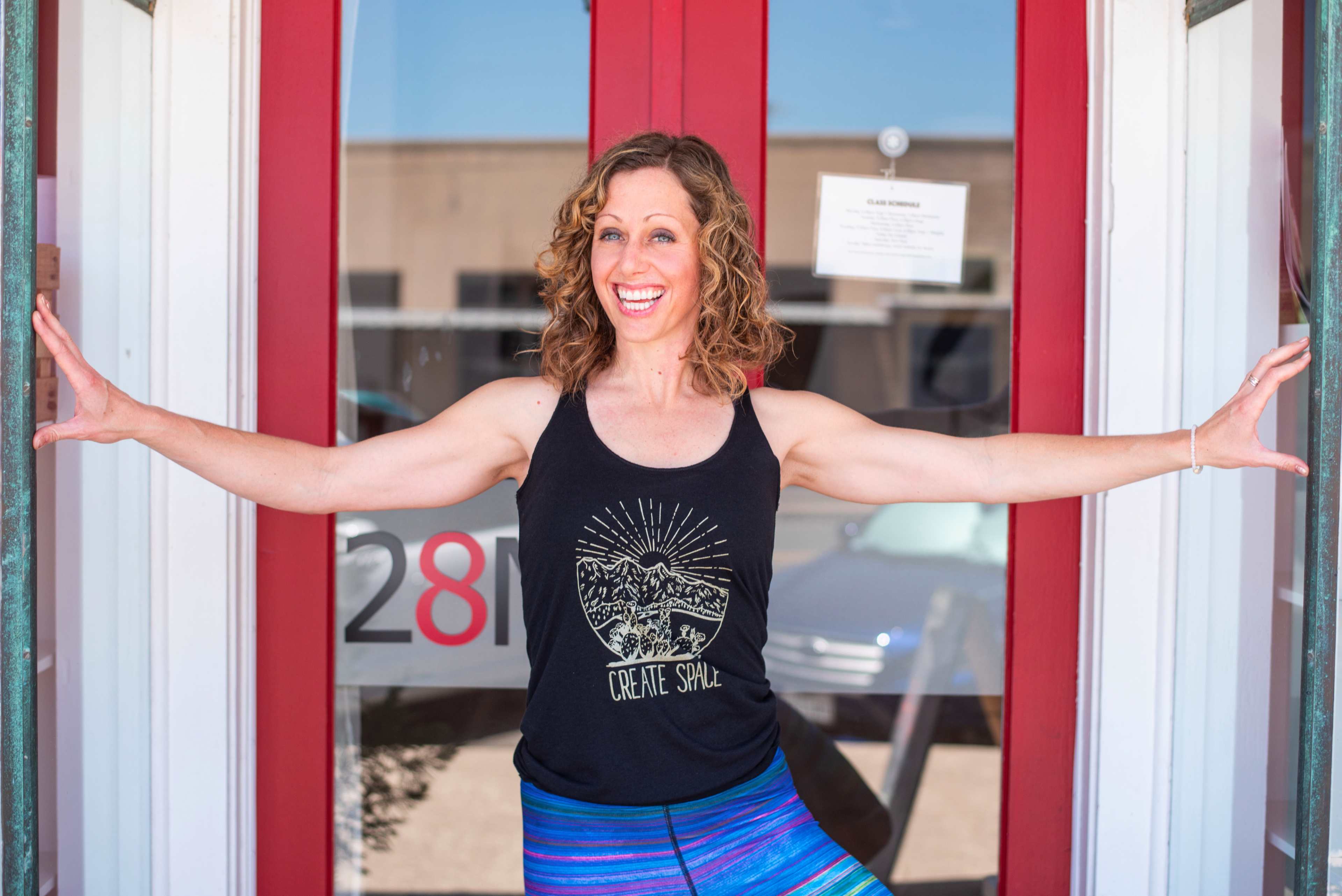 A woman with curly hair stands in front of a red entrance, smiling and posing with her arms extended.