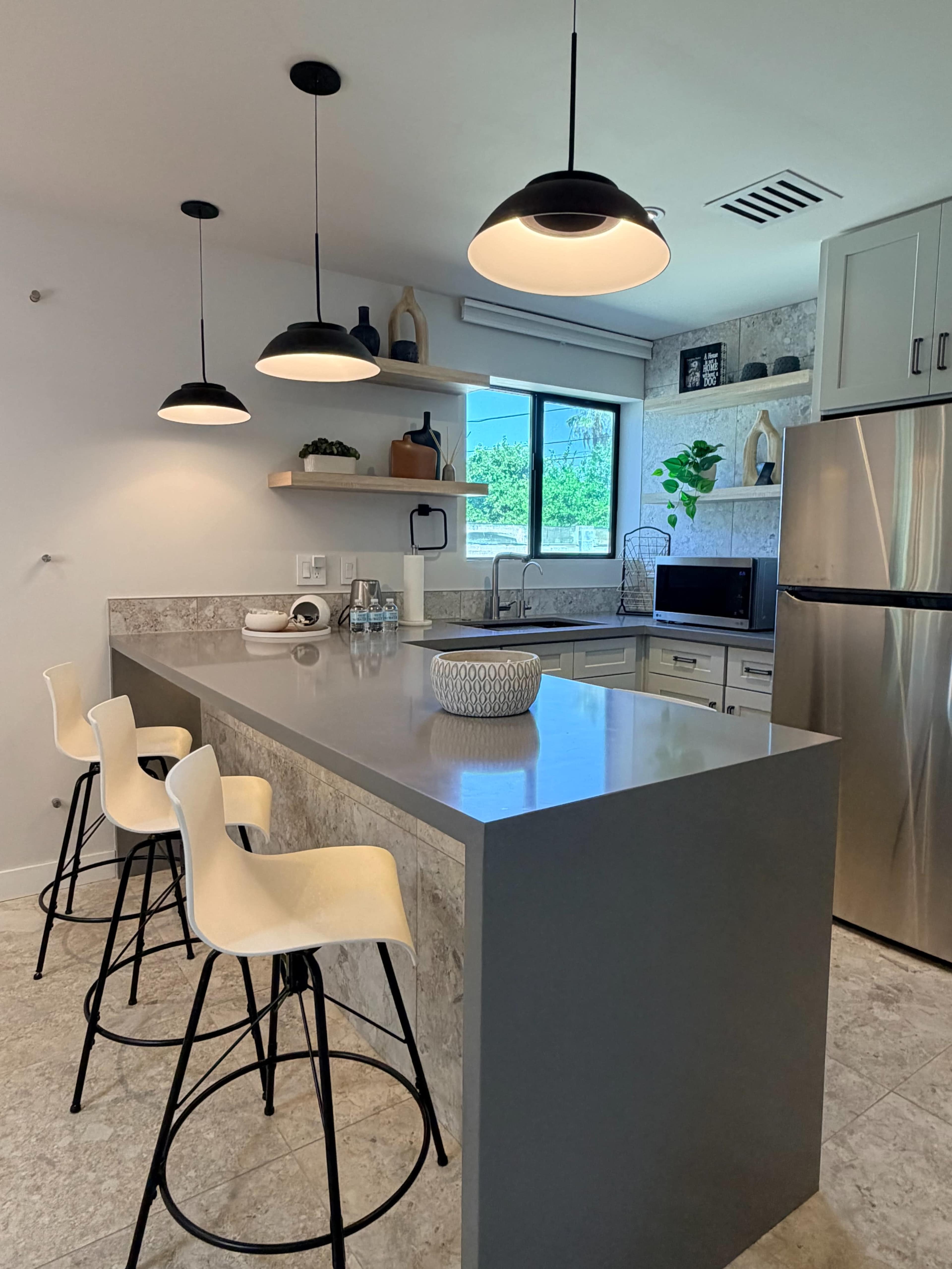 The image shows a modern kitchen featuring a gray countertop with three bar stools, pendant lighting, and shelves displaying decorative items.
