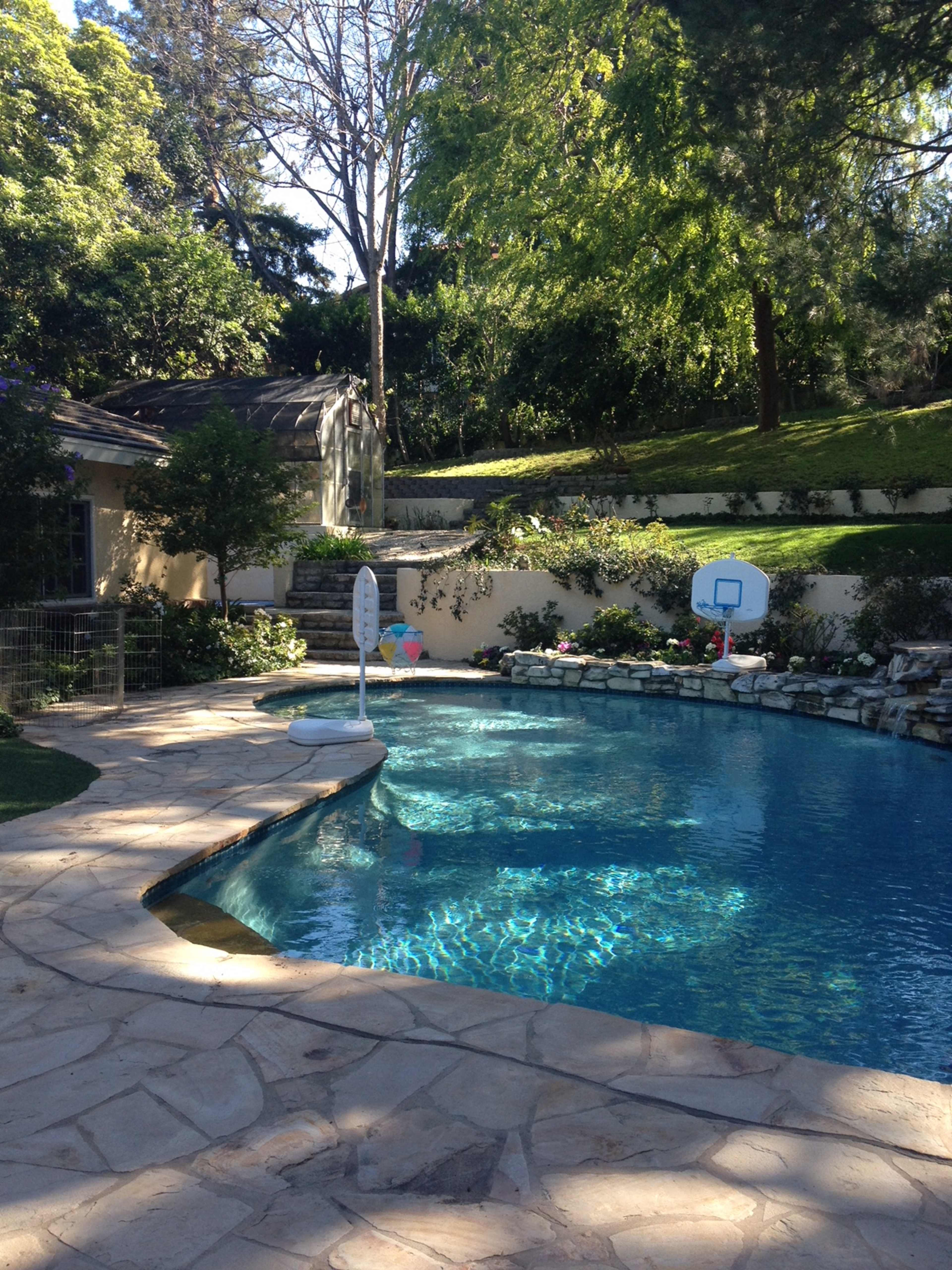 A swimming pool with a stone patio, set in a landscaped yard featuring trees and a basketball hoop.