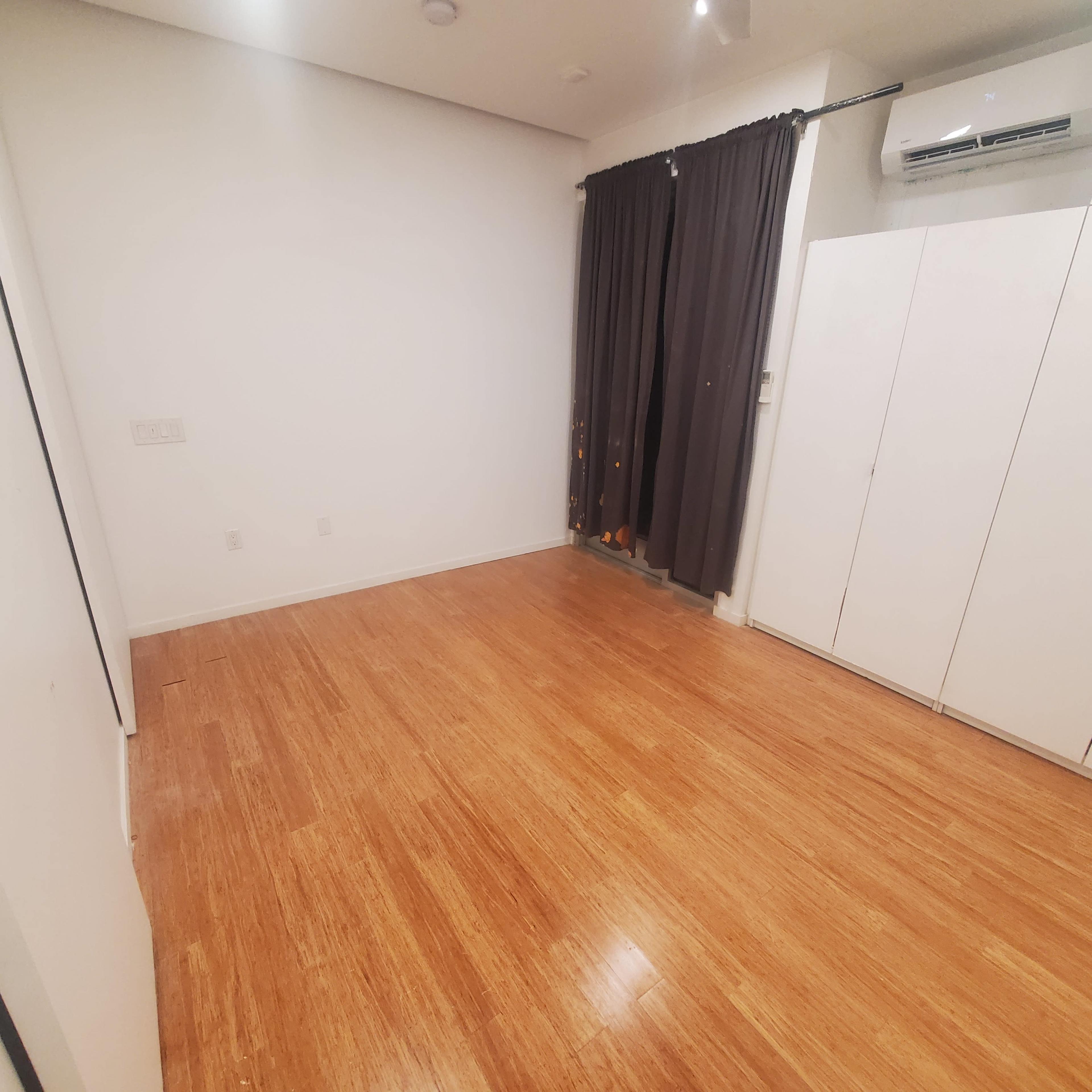 The image shows a sparsely furnished room with bamboo flooring, white walls, and a set of closed cabinets next to a window covered by dark curtains.