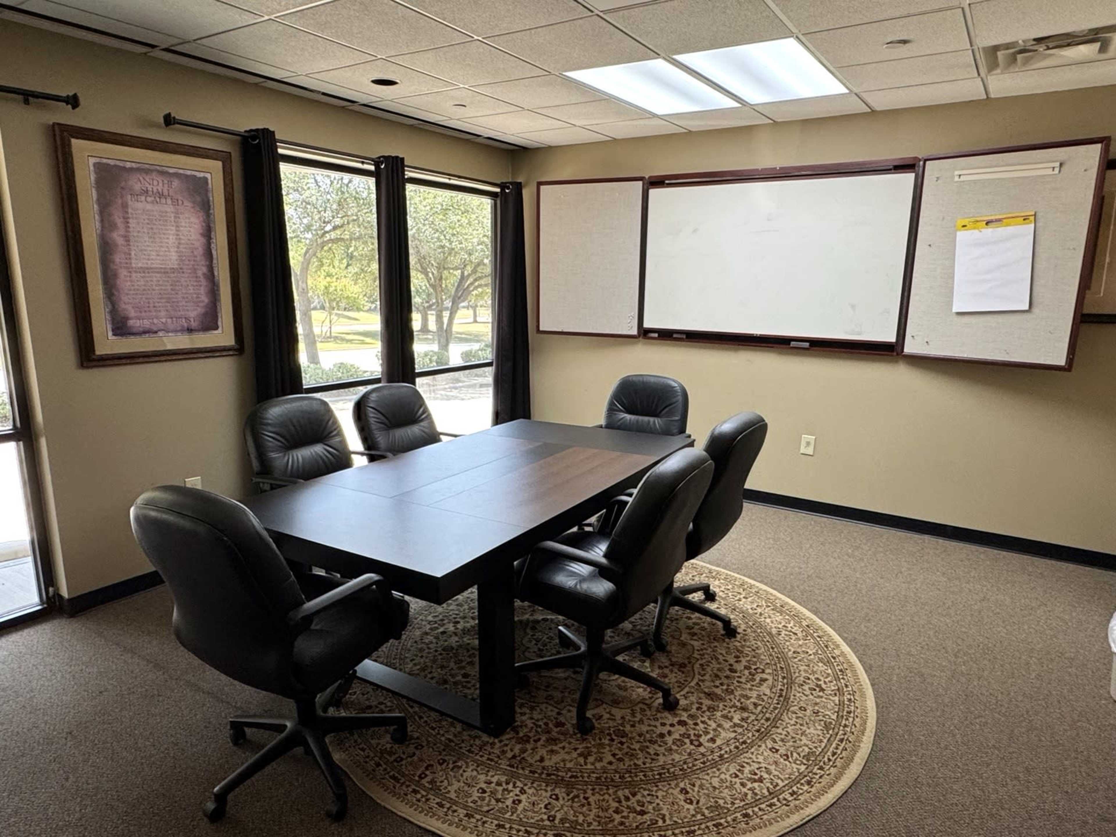 The image shows a conference room with a large table surrounded by six black leather chairs, featuring windows that provide natural light and a whiteboard on the wall.