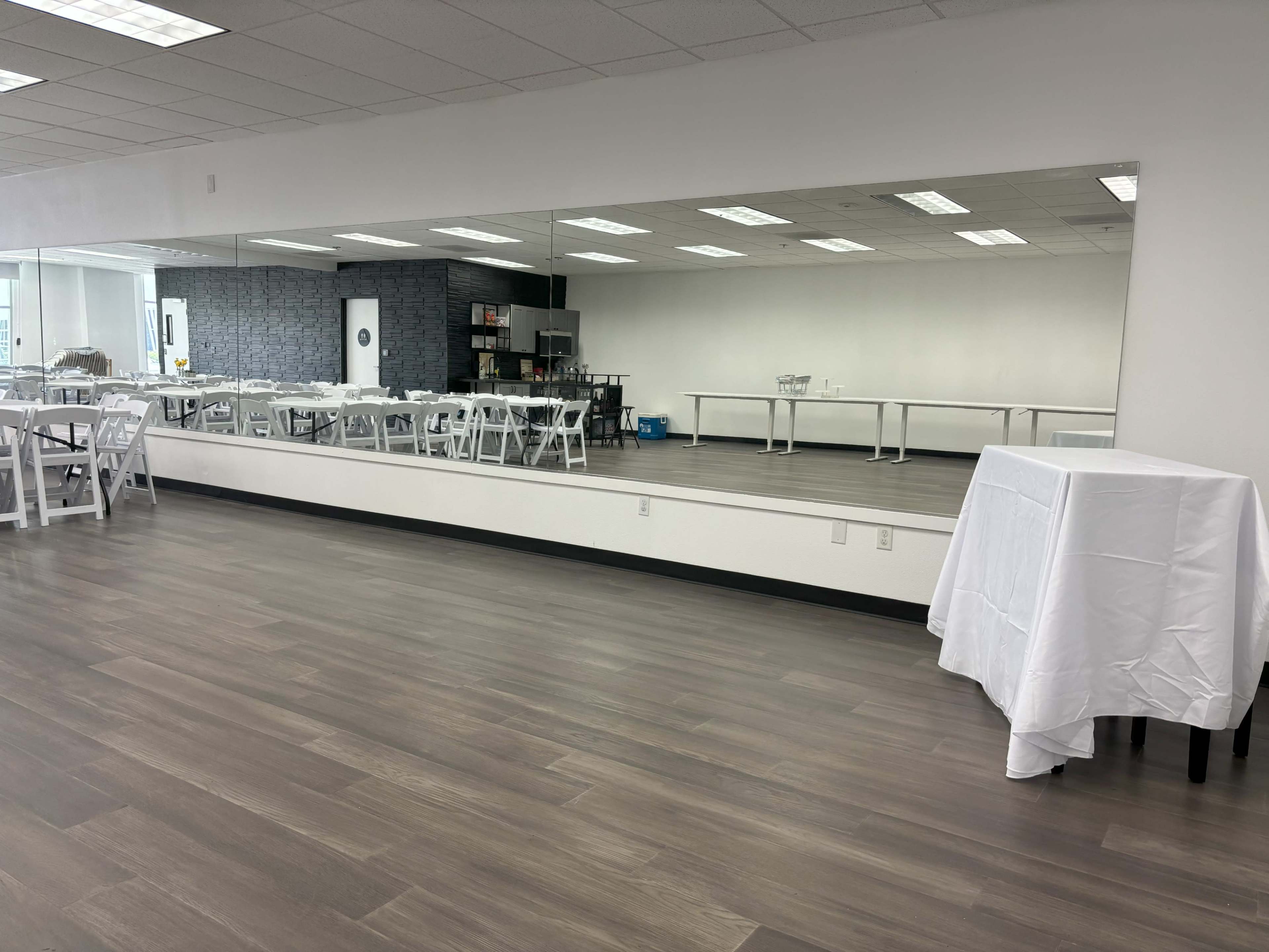 A spacious room with white tables and chairs arranged neatly, featuring a large mirror along one wall.